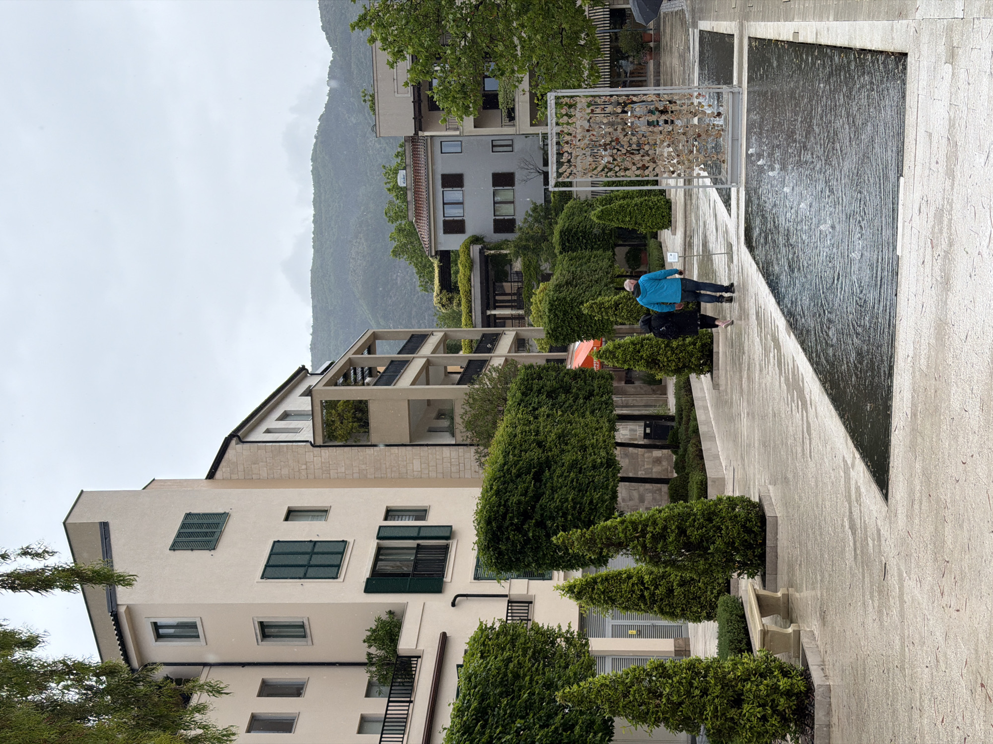 The image depicts a street in Tivat, Montenegro on an overcast day. In the foreground is a sidewalk with a rectangular water feature to the right. To the left are manicured trees, shrubs and benches along with light colored buildings with green shutters. In the background, mountains are visible through the buildings. Two people are walking along the sidewalk in the direction of the water feature. The person in front is wearing a blue jacket, and the person behind is wearing a dark colored jacket or coat. There is also a small, modern sculpture made of circular metallic pieces in the background.