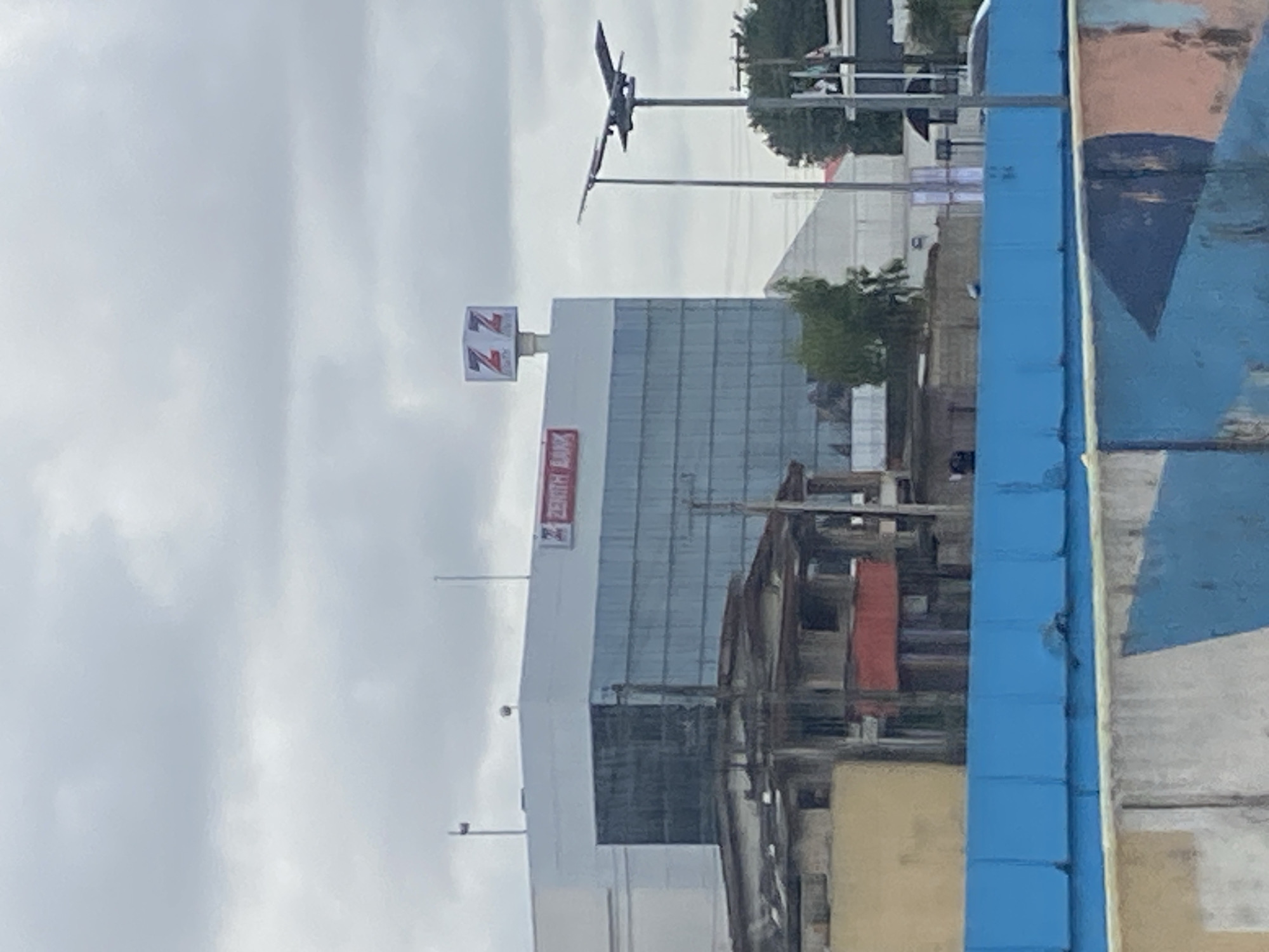 The image captures a cloudy day in Ikeja, Nigeria, focusing on a modern Zenith Bank building. The building features a glass facade and a prominent sign with the Zenith Bank logo on its rooftop. To the left of the bank, older, more dilapidated structures are visible, with weathered walls and exposed roofing. In the foreground, a bright blue barrier runs horizontally across the image, partially obscuring a concrete wall with abstract painted designs. A lamppost with an artistic sculpture of a bird in flight stands to the right of the bank, silhouetted against the sky. The overall scene presents a contrast between modern development and older infrastructure in an urban Nigerian setting.