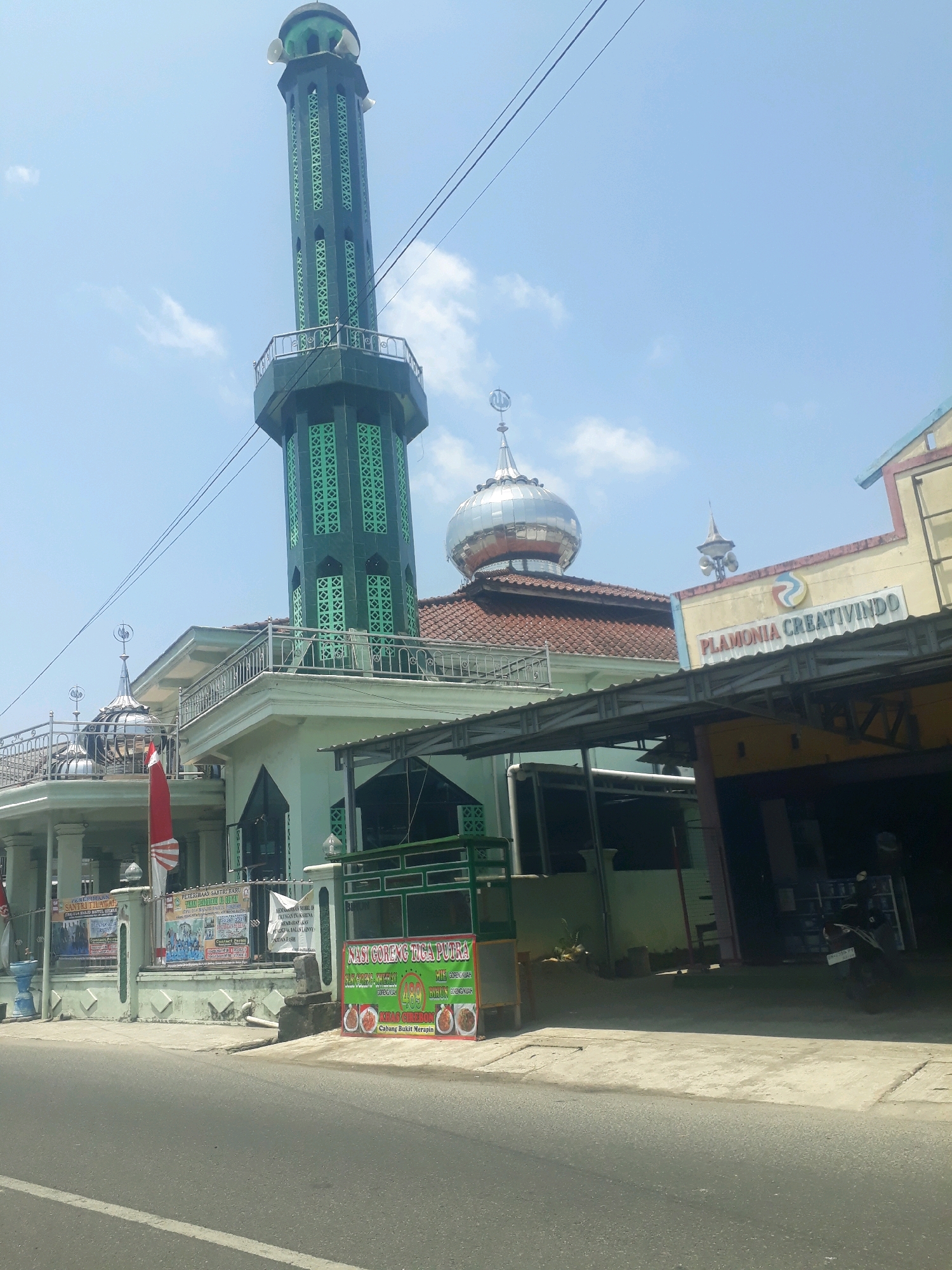 The image shows a mosque with a tall, slender minaret that is dark green with intricate geometric patterns in a lighter green. The mosque has a domed roof, and several smaller domes with crescent moon finials. A red and white Indonesian flag is visible near the entrance of the mosque. In front of the mosque, there are several banners and signs, including one advertising "NASI GORENG TIGA PUTRA". To the right of the mosque, there is a building with a yellow facade and a sign that reads "PLAMONIA CREATIVINDO". The scene appears to be on a street in Pangkalpinang, Indonesia, under a clear blue sky with a few scattered clouds. The street is paved, and there is a sidewalk in front of the buildings. A motorcycle is parked on the sidewalk to the right of the frame. The overall impression is of a sunny day in a commercial or mixed-use area of a city.