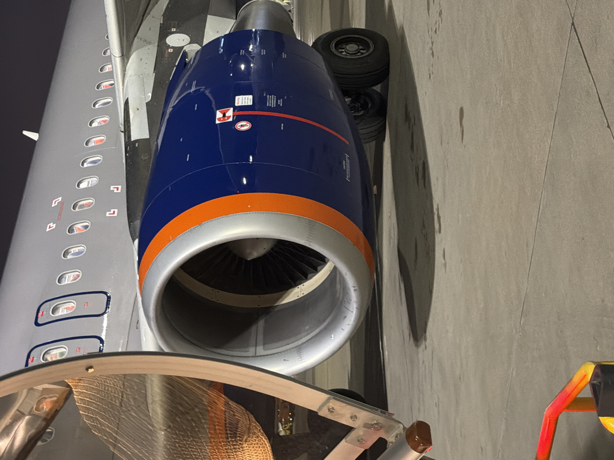 The image shows a close-up, low-angle view of a jet engine on the tarmac at Pulkovo Airport in Saint Petersburg, Russia. The engine cowling is painted navy blue with an orange stripe. The aircraft's fuselage, painted grey, is visible above the engine, with rows of windows. A section of an orange and yellow barrier is in the lower left foreground, and a net-covered object is partially visible to the left of the engine. The ground is concrete tarmac with some dark stains. The lighting suggests it is nighttime or a very overcast day.