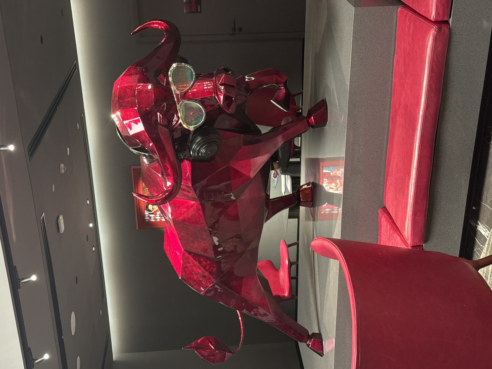 This image, taken in Đà Nẵng, Vietnam, showcases a modern indoor setting, likely a lobby, lounge, or restaurant, characterized by a striking color palette of deep red and dark grey/black. The central subject is a large, vibrant red sculpture of a bull or ox, rendered in a distinctive faceted, geometric style with a highly polished, reflective finish. The sculpture is depicted wearing a pair of light-colored aviator-style sunglasses on its snout, adding a quirky and contemporary touch.
The bull sculpture is positioned amidst matching red upholstered seating, including chairs and possibly a bench or booth, which complement its bold color. These seating arrangements are set against dark grey or black tables and wall panels. The background features a dark wall with subtle recessed spotlights and a linear light strip, creating a well-lit but atmospheric environment. A framed picture with a colorful scene is visible on the wall behind the sculpture. Further back, a dark door is partially visible. One of the wall panels has a small white rectangular device and a faint, almost vertical, light strip. On another wall, near a doorway, the text "PROMO OFFICE" is visible. The scene is devoid of people, presenting a static view of this stylish interior.