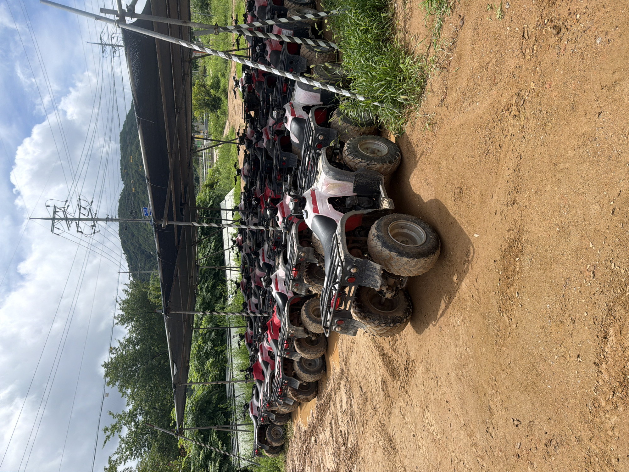 The image shows a large outdoor parking area filled with red and white ATVs, parked under a covered shelter. The ground is made of dirt and appears to be somewhat muddy, with tire tracks visible. In the background, there are trees and hills, with power lines and utility poles also present. The sky is partly cloudy. The ATVs are lined up neatly in rows, suggesting they are available for rent or as part of an organized activity. The scene is likely located in a rural or semi-rural area, possibly a recreational park or a rental facility. The location is identified as Seo-myeon, South Korea.