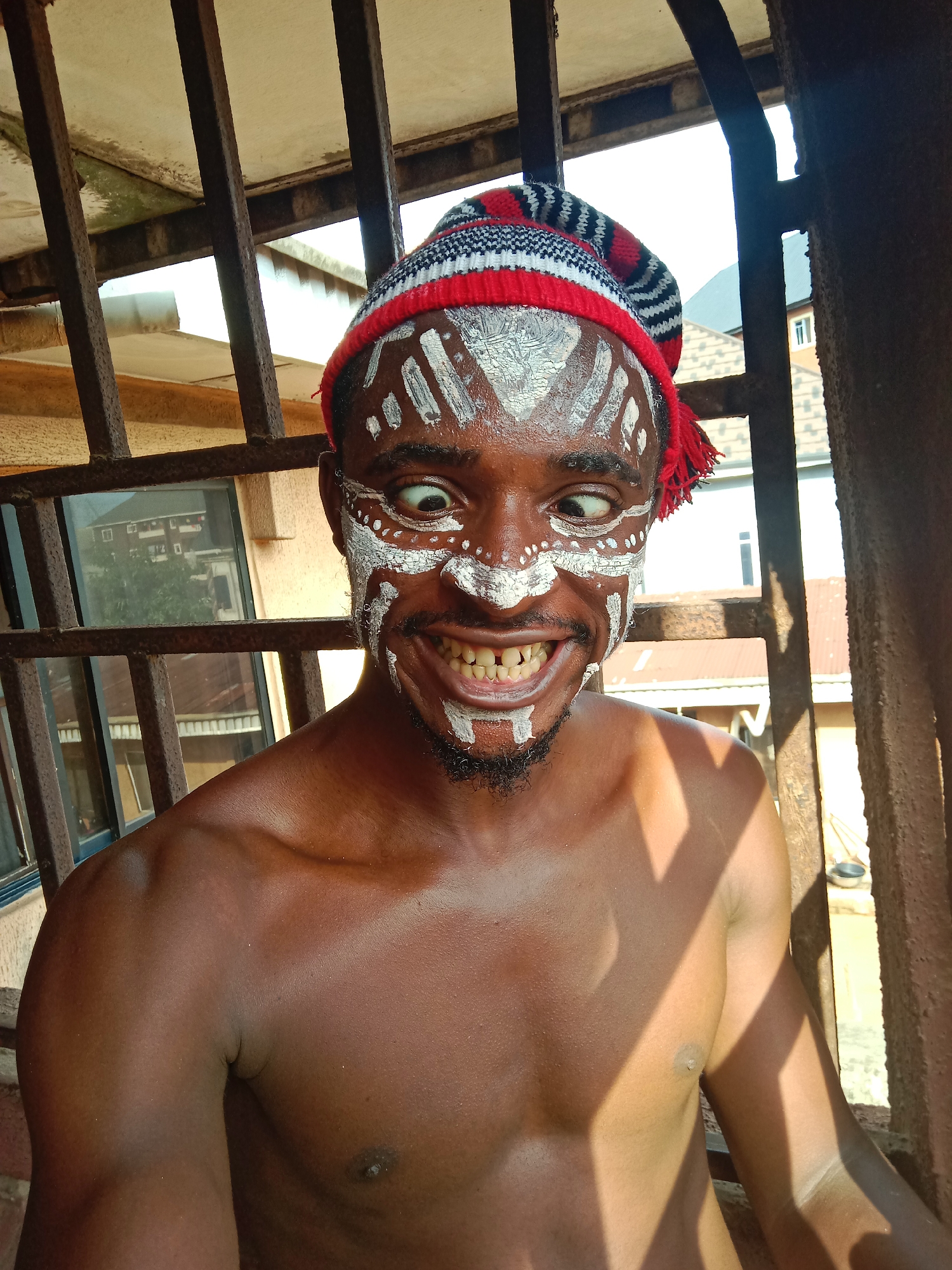 A shirtless dark-skinned man with white tribal-like paint on his face, wearing a red, white, and black striped hat, is standing behind a barred window or gate. He is cross-eyed, grinning, and looking towards the right. The location is likely Nkpor, Nigeria, as indicated in the prompt. Buildings can be seen through the bars in the background.