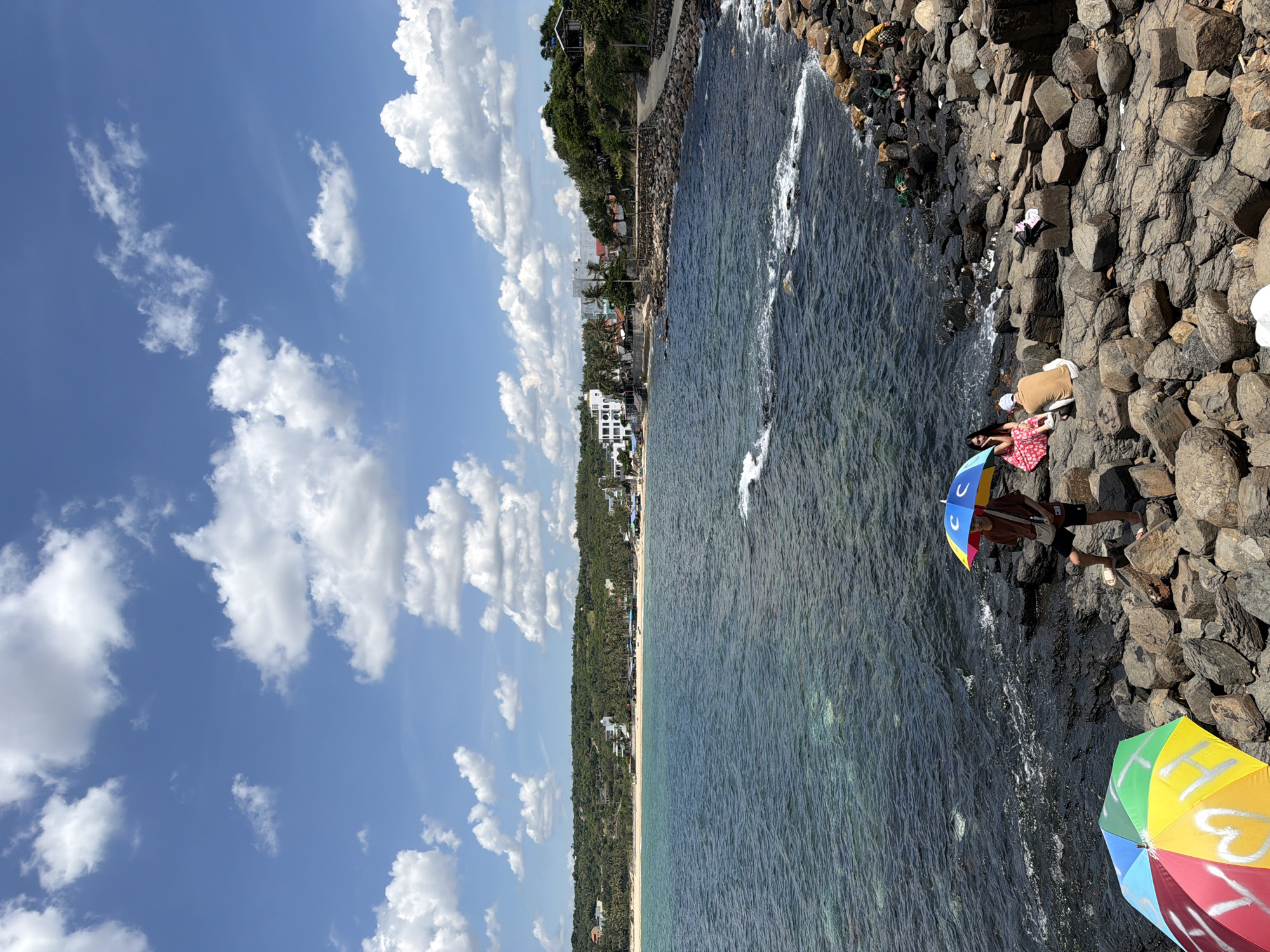 In the city of Phú Lương, Vietnam, this image depicts a scenic coastal view on a bright, sunny day. The sky is a vibrant blue, dotted with fluffy white clouds. The foreground is dominated by a rocky shoreline, with dark, irregularly shaped stones creating a rugged texture. Several people are present, enjoying the beachside. In the lower left, a colorful umbrella, predominantly yellow, green, blue, and red, is partially visible. Further into the scene, a person stands near a rocky outcrop, holding a closed umbrella that is blue and yellow with rainbow accents. Two individuals are seated on the rocks in the middle ground, one wearing a red and white patterned dress and the other in a tan shirt and white cap. A small, white object is placed on a rock between them. To the right, further along the rocky coast, a person in a yellow hat is sitting, and other individuals are visible in the distance. The sea is a deep blue, with clear water near the shore revealing the seabed. In the background, a sandy beach stretches along the coastline, backed by a dense line of green trees and vegetation. A few buildings, appearing to be houses or small establishments, are nestled among the trees. The overall impression is a lively, recreational scene on a warm, clear day at the beach.