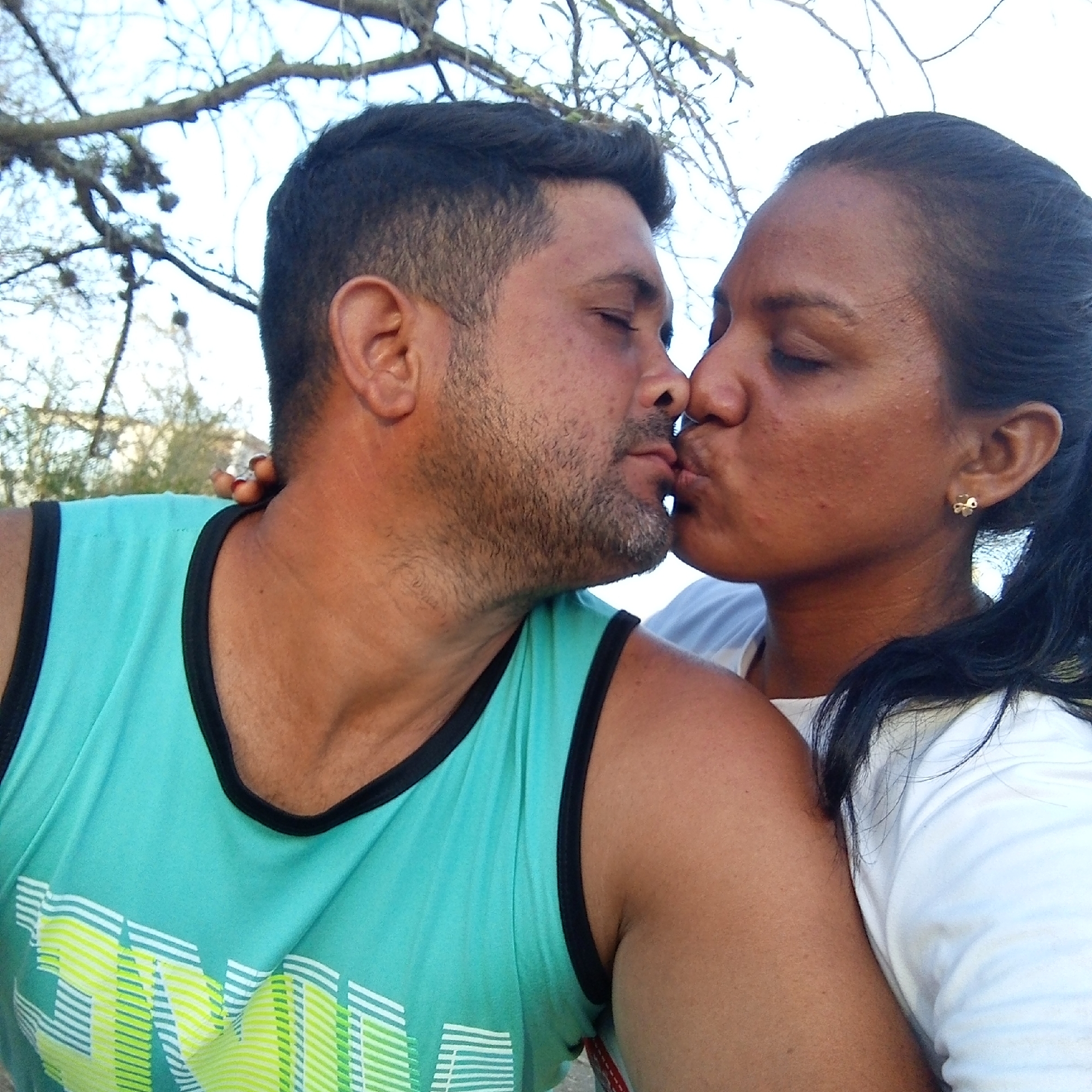 A close-up shot depicts a man and a woman in an intimate embrace, their lips touching in a kiss. The man is wearing a teal tank top with black trim and a graphic design in shades of yellow and white. His hair is short and dark, and he has a beard. The woman has dark hair pulled back in a ponytail and is wearing a white shirt. She has a small gold-colored earring in her ear. The background consists of a sky visible through the branches of a tree. The location is in the city of Unknown, Unknown.