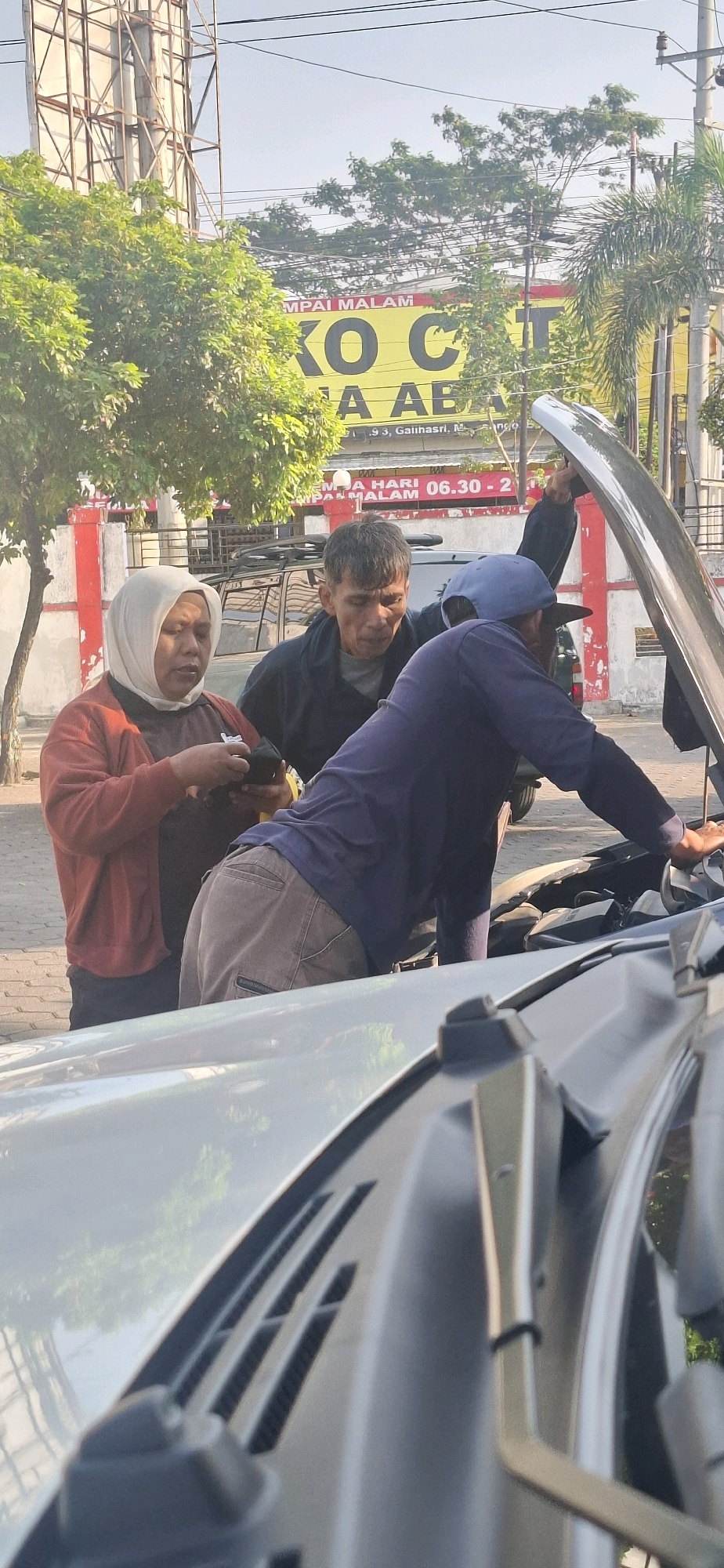 Three people are gathered around the open hood of a car. A woman wearing a headscarf and an orange cardigan is in the foreground on the left, looking down at something in her hands. To her right, a man in a dark jacket is leaning forward, looking into the engine bay. A third person, wearing a blue baseball cap and a navy blue long-sleeved shirt, is also looking into the engine. The scene appears to be a roadside in Gumulan, Indonesia, as indicated by a partial sign in the background that reads "293, Galihasri, M". The environment is outdoors with trees and a building visible.