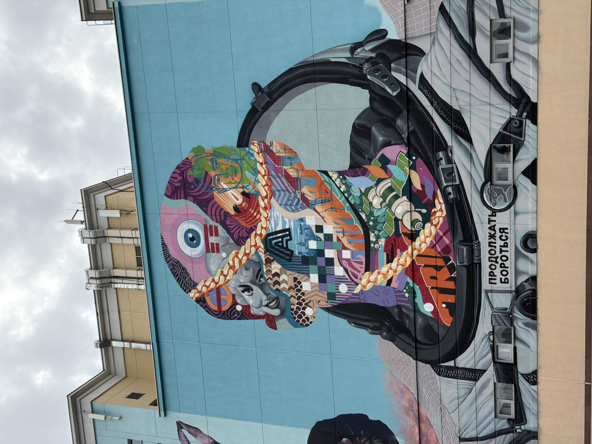 A vibrant, large-scale mural covers the side of a light-blue building under a cloudy sky. The mural depicts an astronaut or cosmonaut figure, with a detailed black helmet and a white and black padded suit, appearing to be in an upright position (despite the image's 90-degree rotation).
The most striking feature is the head within the helmet, which is a complex and colorful collage of diverse artistic elements. This collage includes a large stylized eye, a grayscale portrait resembling Marilyn Monroe with a subtle smile, intertwined golden chains, various geometric patterns (like checkerboards and chevrons), organic shapes, and abstract designs in a multitude of colors. Visible text within the collage includes "INTERKANN" near the eye.
On the white part of the suit, near the right shoulder, Cyrillic text reads "ПРОДОЛЖАТЬ БОРОТЬСЯ," which translates to "CONTINUE TO FIGHT" or "KEEP FIGHTING." Several small windows from the actual building are integrated into the mural on the suit's design, creating a trompe l'oeil effect.
The background shows the light blue wall of the building, with other building structures and a heavily overcast sky visible towards the top and left of the frame. The image captures daytime conditions in an urban setting, likely Moscow, Russia.