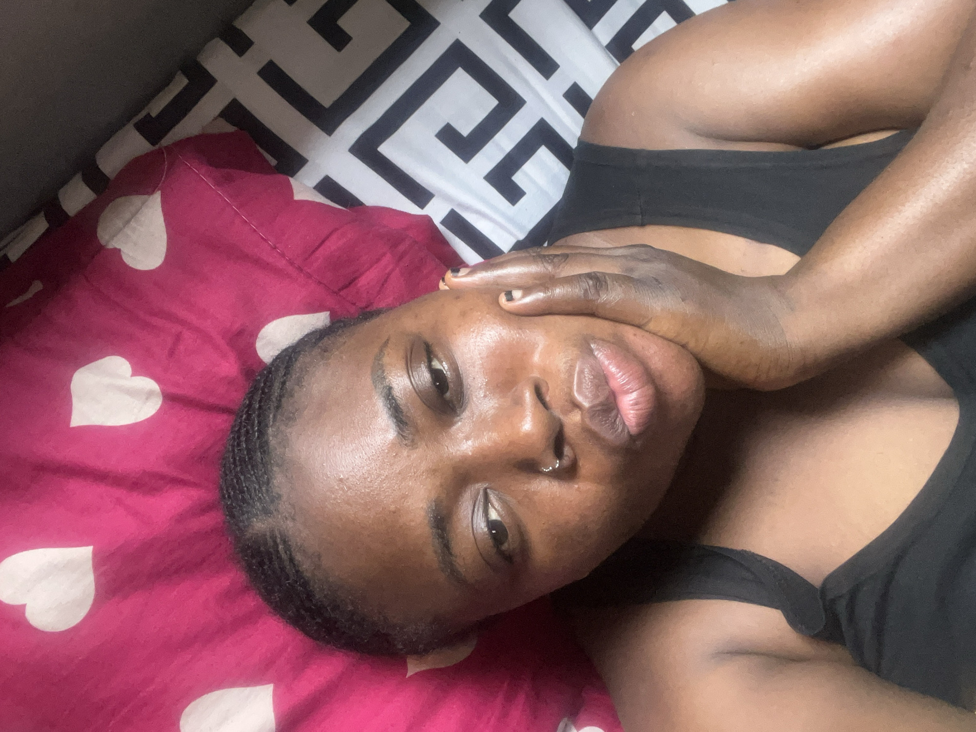 The image is a close-up selfie of a dark-skinned woman lying down. She is on a bed with a red and white heart-patterned pillow. She's wearing a black tank top. Her hair is styled in braids. Her right hand is holding her face. The location is likely a bedroom, but there is no visual information suggesting it is in Abuja, Nigeria.