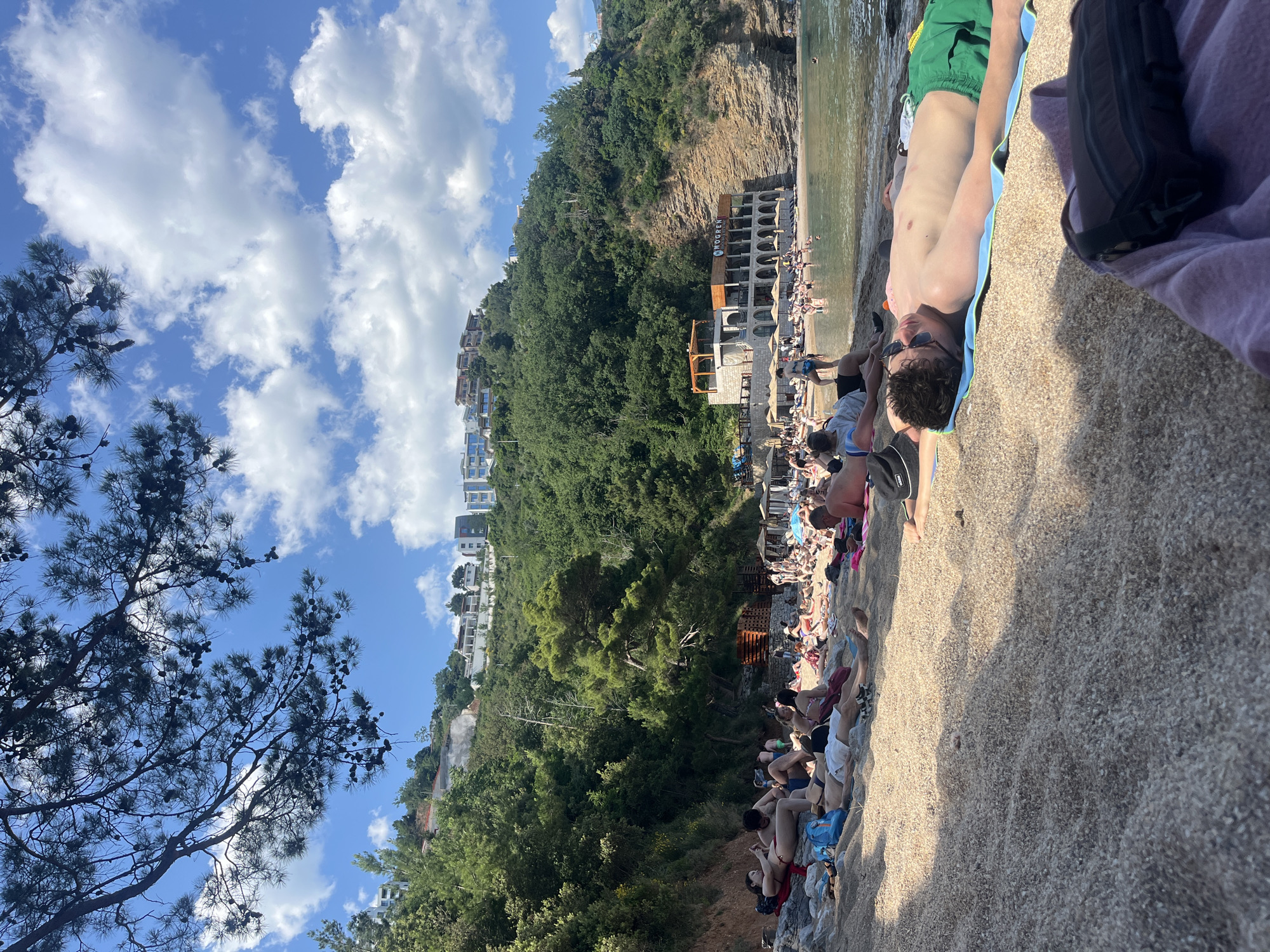 The image shows a beach scene in Budva, Montenegro. A crowded beach stretches along the coast with a building near the water. People are relaxing, sunbathing, and swimming. A steep, green hillside rises behind the beach with buildings perched along the slope. The sky is blue with white clouds. A person wearing sunglasses is lying on a towel in the foreground. The photo is taken from ground level looking upwards.