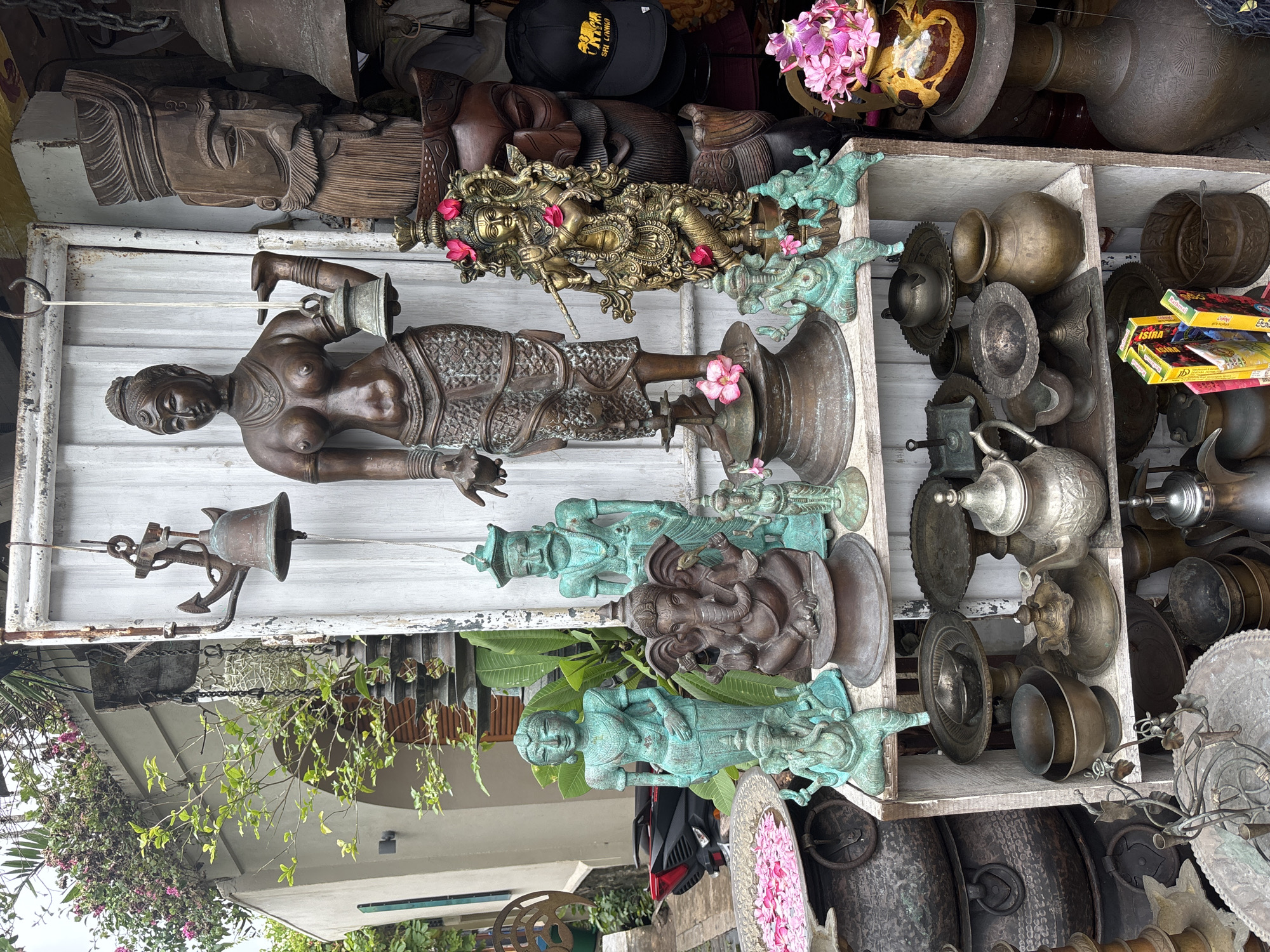 This image captures a roadside display of vintage metal artifacts and religious statuettes in Galle, Sri Lanka. The items are arranged on aged white wooden shelves. The display includes copper and brass pots, various ornate jugs, and figurines of deities. Specifically, there is a dark bronze statue of a bare-breasted woman holding a bell, along with multiple turquoise-patinaed figures, including one of Ganesha. A carved wooden head lies on the top shelf, next to a black cap with the words "SRI LANKA" written on it, as well as a statue of a man with pink flowers woven into it. There are potted plants on the lower left and right corners of the shelves. The foreground shows a glimpse of street activity, and a dark red motorbike is visible in the lower center.
