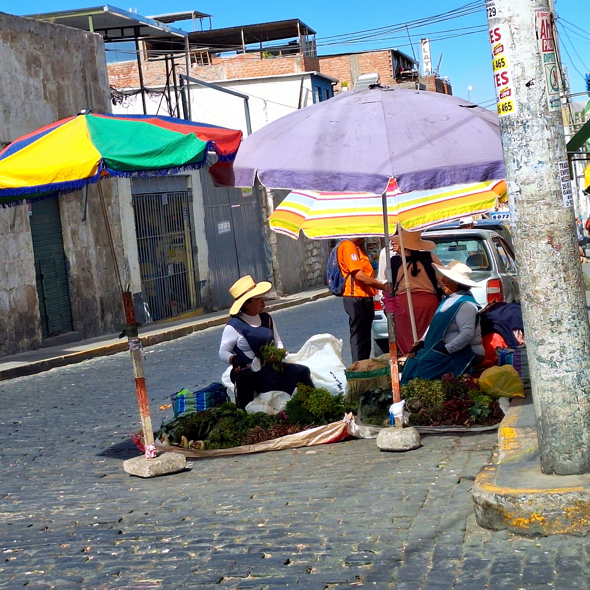 The image shows a street scene in an unknown city. The cobblestone street has two women vendors selling produce under large umbrellas, one with yellow, green and red panels, and the other in light purple. A third person, an individual wearing an orange shirt, stands in the background beside a car. The vendors are seated amongst their merchandise, which includes various greens and vegetables. Buildings line the street, and a utility pole covered in posters stands on the right side. Overhead, electrical wires crisscross the sky.