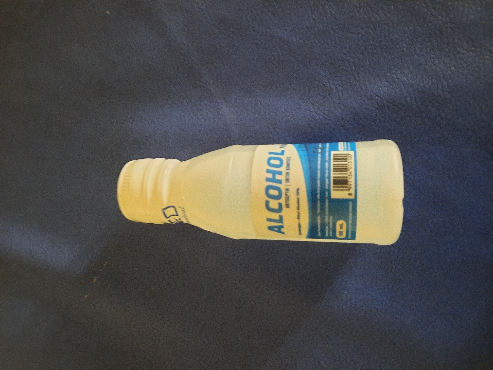 A white plastic bottle containing a clear liquid, labeled "ALCOHOL 70% ANTISEPTIK | UNTUK KOMPRES", is centrally placed on a dark blue textured surface. The bottle has a white screw-top lid. A blue and white label wraps around the lower portion of the bottle, displaying text and a barcode. The bottle's contents appear to fill most of the container. The background is an out-of-focus dark blue textured surface, possibly a fabric or a mat. The lighting suggests a single overhead light source, casting a subtle shadow to the left of the bottle. There are no people visible in the image. The image is taken from a slightly elevated angle, looking down at the bottle.
The image is a product photograph of an antiseptic alcohol bottle. The context of Lhokseumawe, Indonesia is not visually represented in the image.