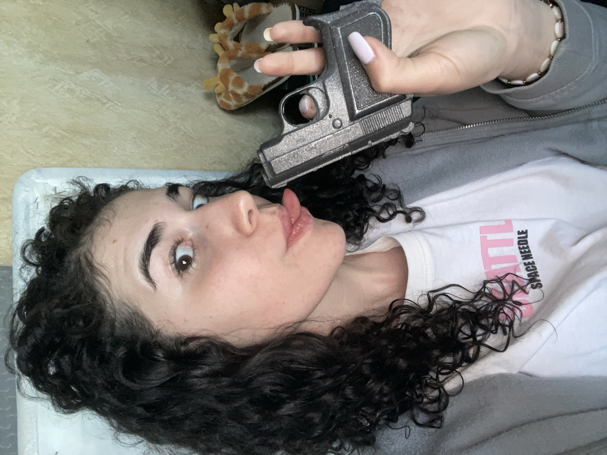 A woman with dark, curly hair lies on a patterned surface, possibly a bed, with her head tilted back. Her mouth is open, tongue extended toward a toy gun held by another person above her head. The hand holding the gun has painted nails and wears a bracelet, and the arm is covered by a grey hoodie. Part of a stuffed animal is visible in the upper left corner of the image. The woman is wearing a white t-shirt with the words "SPACE NEEDLE" in pink and black lettering. The location is unknown and could not be determined based on the information provided in the image.