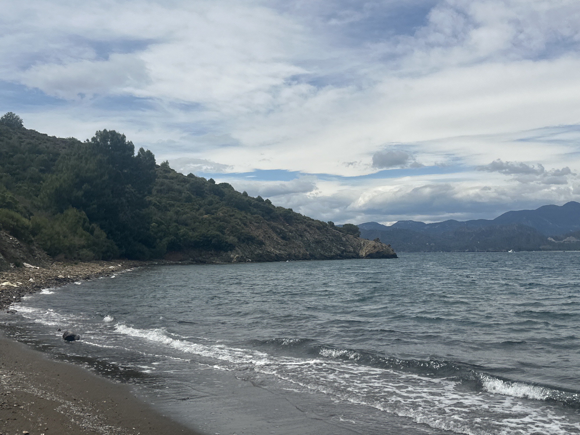The image shows a shoreline along a bay with mountains visible in the distance under a cloudy sky. The foreground features a dark sandy beach with gentle waves washing ashore. A rocky headland covered with trees extends into the bay from the left side of the frame. The water is a dark shade of blue-grey, reflecting the overcast sky. A small boat is visible in the distance near the mountains. The location is likely along the coast of Fethiye, Turkey, based on the region's geography.
