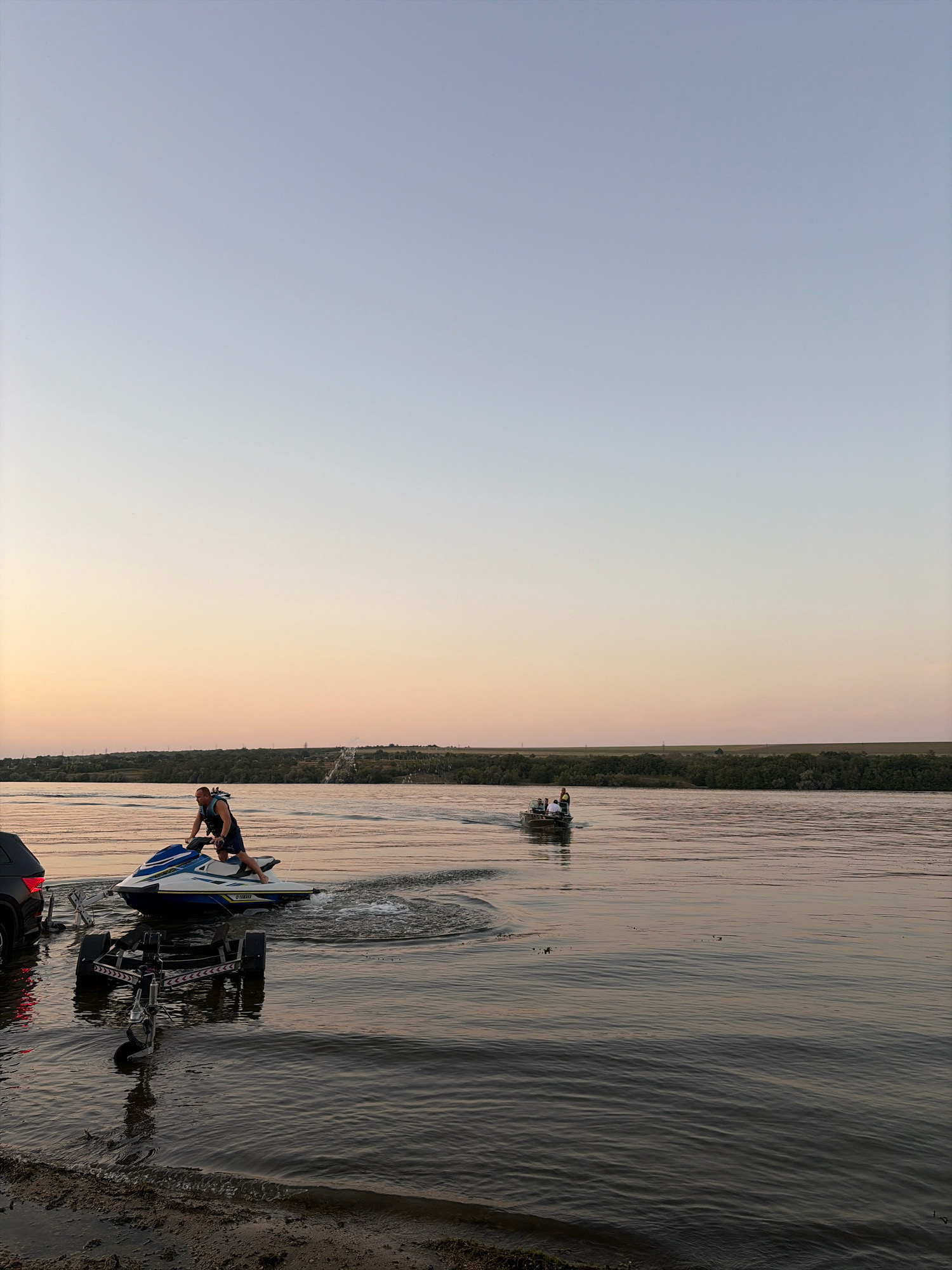 A man on a jet ski is launching it into a body of water, presumably a river, as part of a boat trailer is submerged in the water next to it. The man is wearing a life vest and appears to be in motion. In the background, a small boat with several people is moving away from the shore. The environment is a calm body of water with a tree-lined bank in the distance. The sky is clear with a gradient of light blue and soft orange hues, suggesting it is either sunrise or sunset. The location is identified as the city of Oxentea, Moldova.