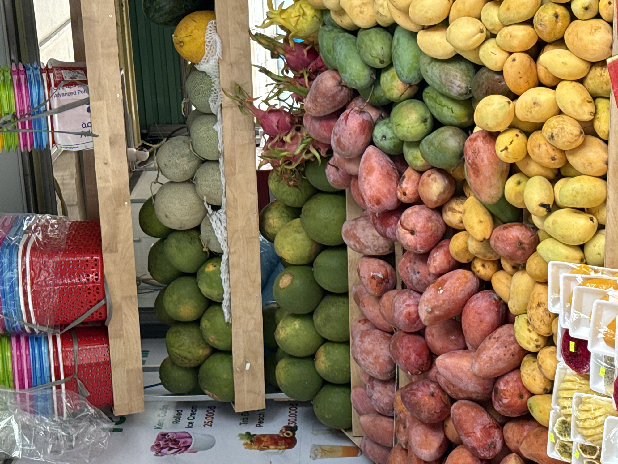 The image depicts a colorful fruit stand in Nha Trang Ward, Vietnam. The stand is stacked with a variety of fruits, including melons, dragon fruit, green mangoes, and ripe yellow mangoes. Other items visible include stacks of plastic baskets, a bag of a product labeled "Advanced Pet" and advertising for "Kem Cuôn Rolled Ice Cream" and "Trà Đ Peach" priced at 25,000₫ and 30,000₫ respectively. The fruits are arranged in vertical columns, showcasing the assortment of produce available. The overall scene appears to capture a typical fruit vending location in the ward.