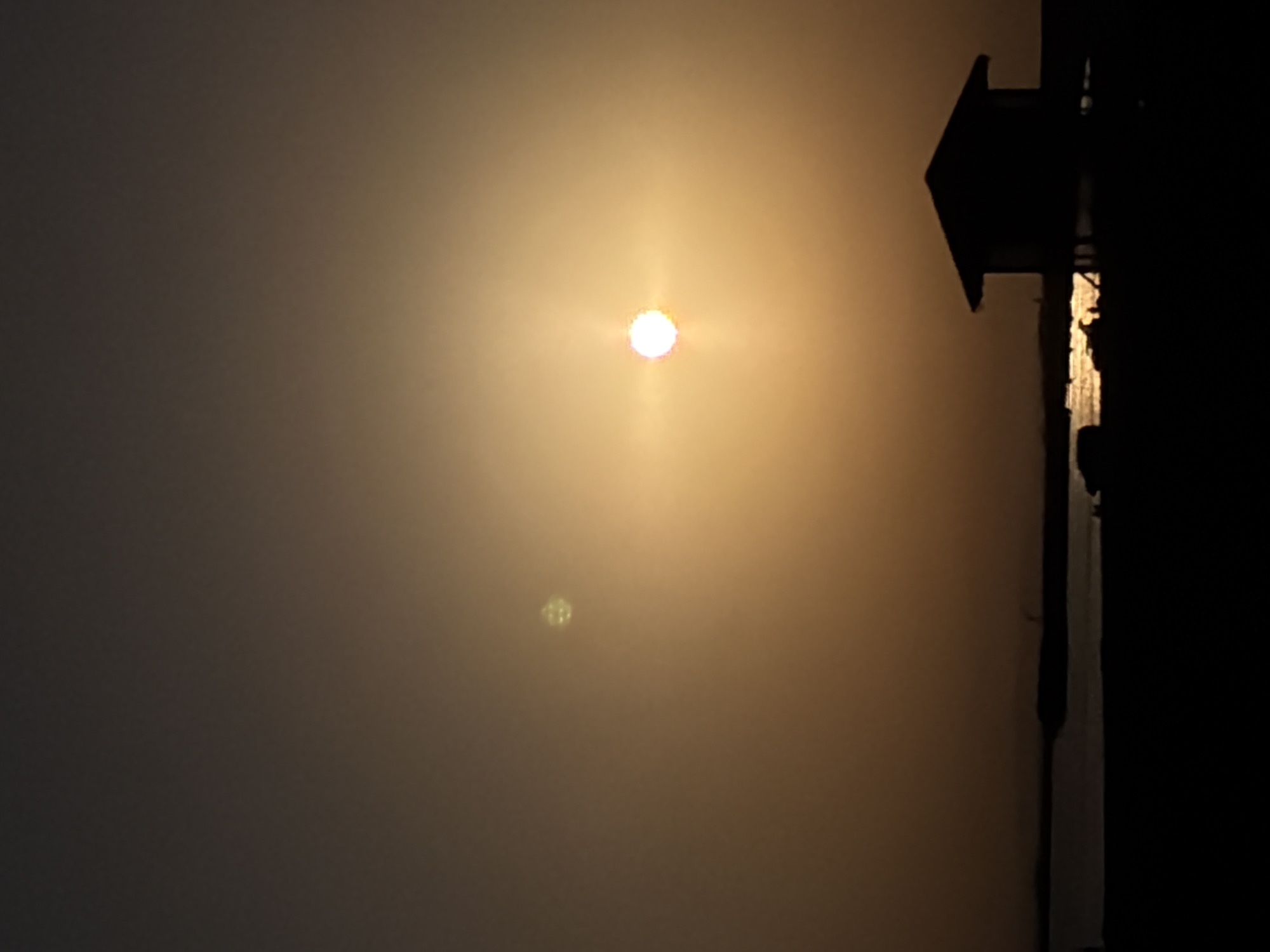 The image depicts a sunset over the Caspian Sea in the Pirallahi Raion of Azerbaijan. The sun is a bright, diffused orb in the hazy sky, casting a warm orange glow. A small, silhouetted structure, resembling a beach hut or lifeguard station, stands on stilts in the foreground, facing the water. The water itself reflects the sunlight, creating a shimmering effect. The horizon is low and indistinct, with a darker landmass or distant shore visible. The overall mood is serene and tranquil, characterized by the warm light and the quiet natural setting. There are no people visible in the image.