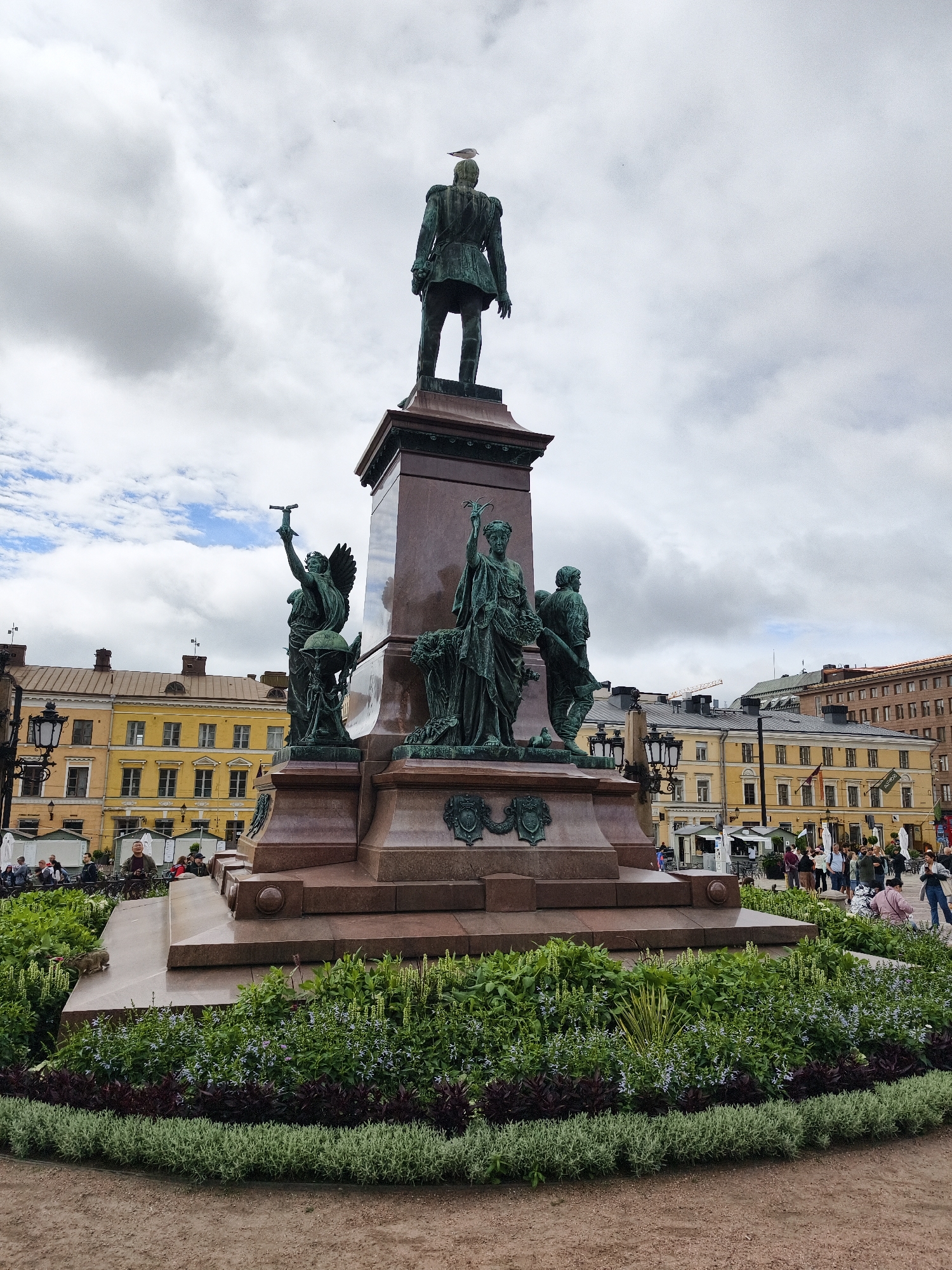 The image shows a statue of Alexander I of Finland in Senate Square in Helsinki, Finland. The statue is made of bronze and is mounted on a granite pedestal. The statue depicts Alexander I standing and wearing a military uniform. There are also four allegorical figures at the base of the pedestal, representing peace, justice, wisdom, and strength.
In the foreground, there is a circular flowerbed with a variety of plants, including green bushes and purple and blue flowers. In the background, there are several yellow buildings with red roofs, which are typical of Helsinki's architecture. People are seen walking around the square, and there are also some tents and market stalls. The sky is cloudy, with some patches of blue sky visible.
The statue of Alexander I is a prominent landmark in Helsinki and a popular tourist attraction. It was erected in 1894 to commemorate the 100th anniversary of his birth. The statue is made of bronze and is about 5 meters tall. The allegorical figures at the base of the pedestal are made of marble.
