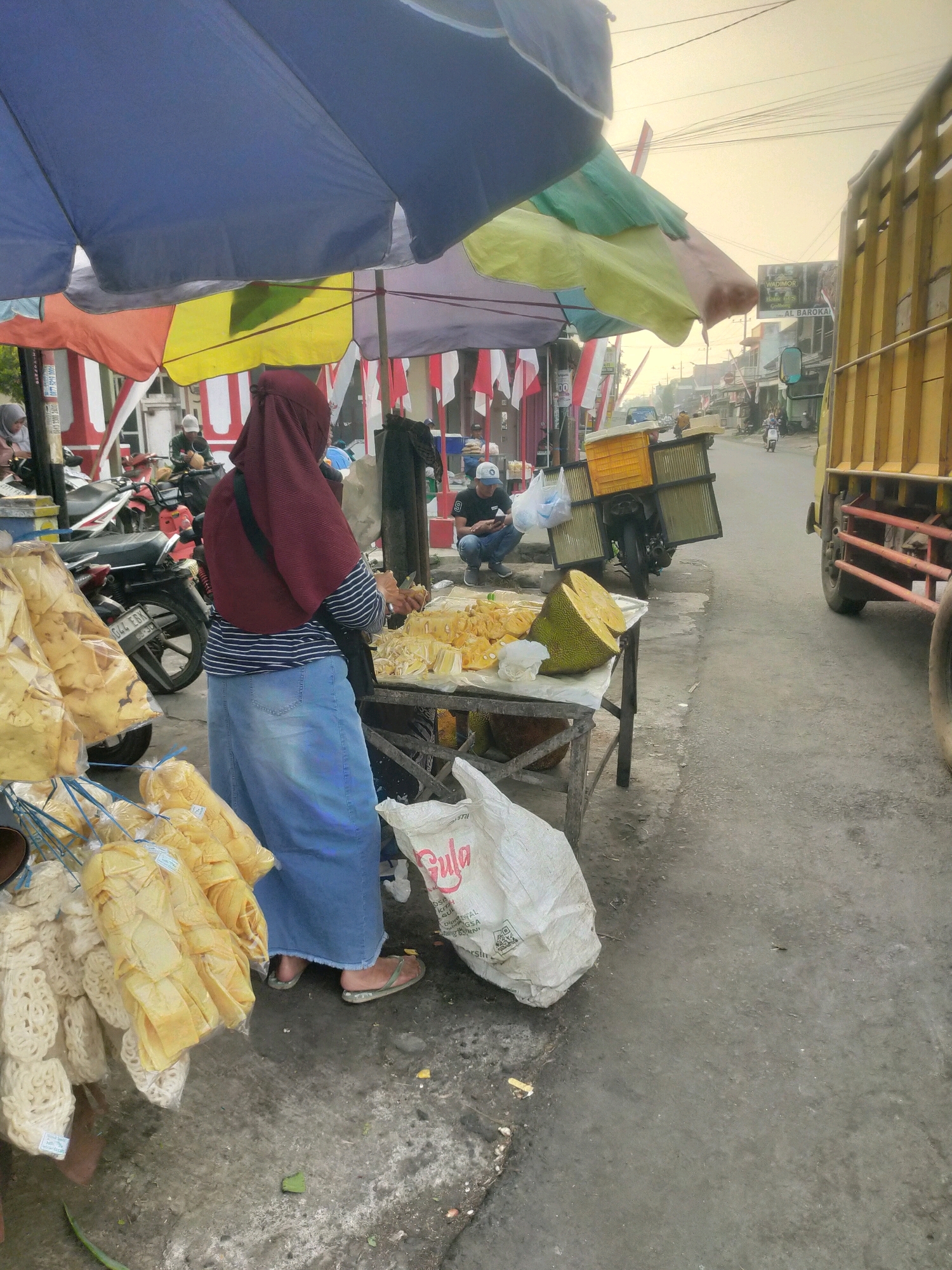 The image depicts an outdoor market scene in Bululawang, Indonesia. A woman wearing a maroon hijab and a long blue denim skirt stands behind a stall, holding a knife and appears to be cutting jackfruit. The stall is laden with cut jackfruit and whole jackfruits. To the left of the stall, several plastic bags filled with yellow chips or snacks are hanging, and a larger bag of white, circular snacks is also visible. A white sack with the word "Gula" (sugar) printed on it lies on the ground near the stall. In the background, a yellow truck is parked on the right side of the road, and a motorbike is parked on the left. Several other people are present in the market, including a person in a white cap sitting on the ground in the middle ground, looking at a phone, and others partially obscured by the market stalls and tarps. The market appears to be set up along a paved road, with buildings and power lines visible in the distance. The sky is overcast, suggesting either early morning or late afternoon light. The overall impression is of a busy, local market day.
