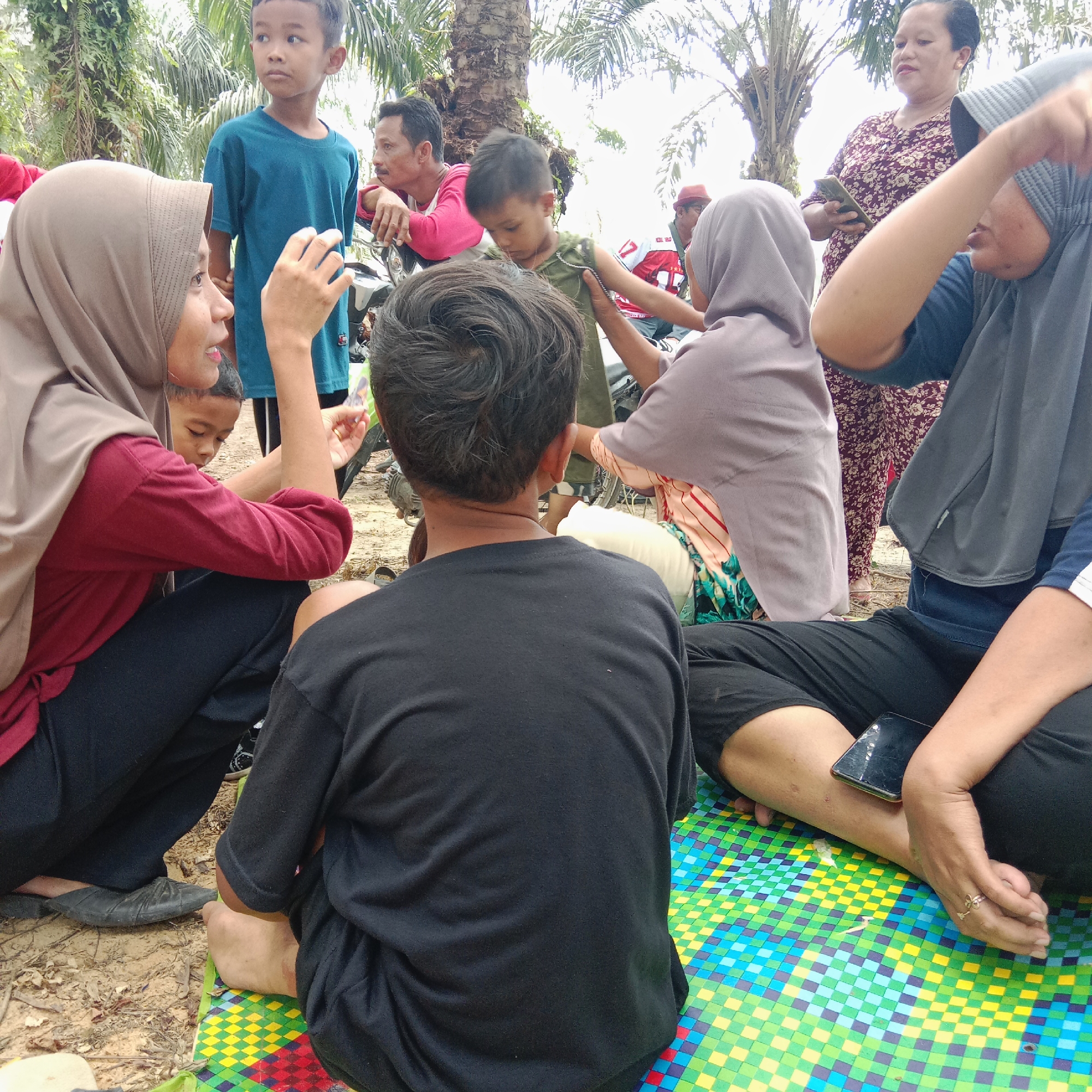 The image depicts a group of people, likely a family, gathered outdoors in what appears to be a rural or semi-rural area. The environment suggests a tropical setting, with palm trees visible in the background. The ground is covered in dry soil and some leaves, and a colorful mat is spread out on the ground where some individuals are seated.
Several people are visible, including adults and children. A woman in a beige hijab and a red shirt is seated on the left, interacting with a young boy. A man in a pink shirt is leaning against a motorcycle in the background. Another young boy in a blue t-shirt stands nearby. A woman in a patterned dress is standing further back, holding a mobile phone. Another woman, wearing a grey hijab, is seated on the right side of the mat. A person in a black t-shirt is seated in the foreground, with their back to the camera.
The overall scene conveys a casual gathering, possibly a picnic or a moment of rest during an outing. The presence of the motorcycle suggests they might have traveled by vehicle. The focus is on the interactions and shared space of the individuals. The image does not provide specific visual cues that would definitively place it within Dumai City, Indonesia, but the environment and the people's attire are consistent with a Southeast Asian context.