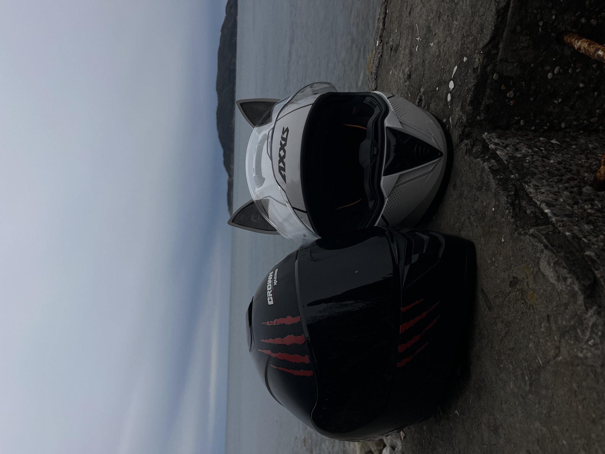 Two motorcycle helmets are lying on a concrete surface near a body of water. The helmet on top is white with gray cat ears and the brand name "Axxis" on the side. The helmet below is black with red claw mark designs. In the background, a gray body of water is visible, with a land mass across the water under a cloudy sky. The location is Kamenovo, Montenegro.