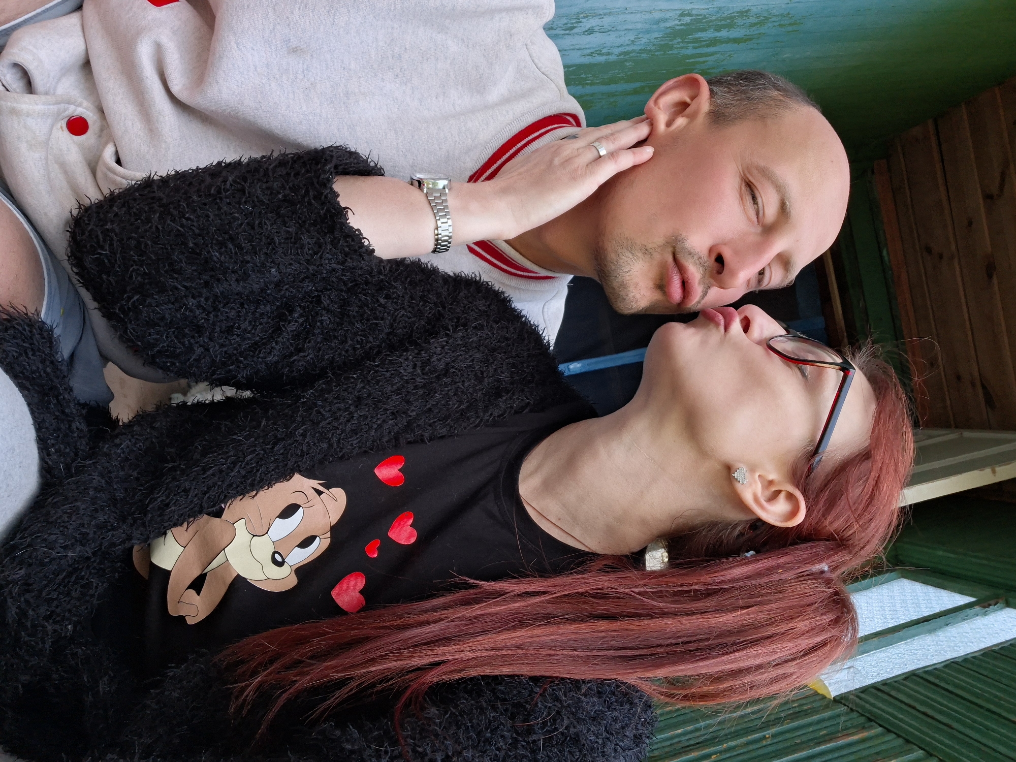 A close-up, selfie-style image shows a man and a woman from the chest up, embracing outdoors, with the man leaning in towards the woman as she kisses him. The woman, on the right, has long, reddish-brown hair tied in a ponytail and wears glasses, a black fuzzy cardigan over a black t-shirt with a cartoon character and red hearts. She also has silver heart-shaped earrings and a silver square earring in her left ear. The man, on the left, is balding and has a mustache and stubble, wearing a light grey sweatshirt with red trim and red buttons. His left hand is on the woman's face, and he wears a silver watch on his left wrist and a silver ring on his left ring finger. Their surroundings appear to be rustic, with a teal-colored wooden wall on the left and a weathered green wooden structure on the right, suggesting an outdoor setting, possibly a porch or an outbuilding. The background shows wooden planks overhead and more green siding. The lighting is natural, indicating daytime. The image is focused on the couple's affectionate interaction.