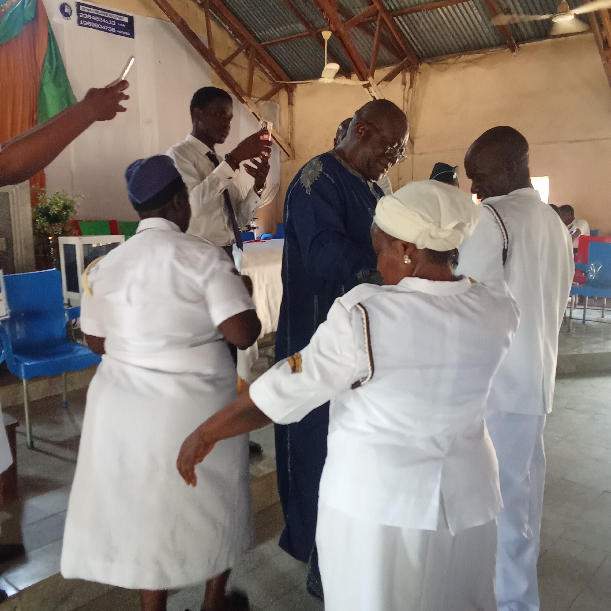 The image captures a scene indoors, likely in a church or a similar gathering space, based on the presence of religious attendants, attire, and background decorations.
Several individuals are present. A man in a dark blue garment is the center of attention. Others dressed in white uniforms that suggest a ceremonial or religious role surround him. A young man in a white shirt and tie is taking a picture of the man in the blue garment.
The environment appears to be a simple, functional space, with corrugated metal roofing and basic furnishings like blue plastic chairs. The background is generally light-colored, with some decorative fabric draping and other symbolic items. The photograph appears to have been taken in Unknown, Unknown.