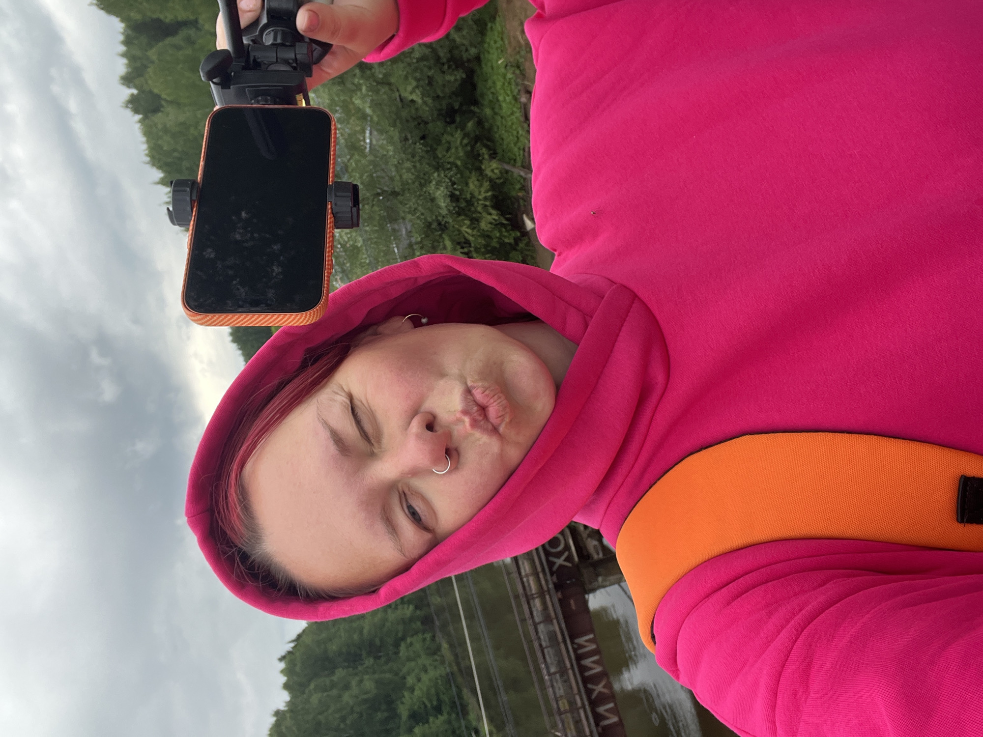 A woman with fair skin, light eyebrows, and a nose ring is taking a selfie. She is wearing a bright pink hooded sweatshirt with an orange strap visible on her shoulder. She is holding a black smartphone in a phone mount.
The background shows a river, trees, and a cloudy sky. Russian text (ИХИИ XO) is visible on a structure near the river. This is likely a location in the city of Chaikovsky, Russia, based on the Russian text and the provided prompt.