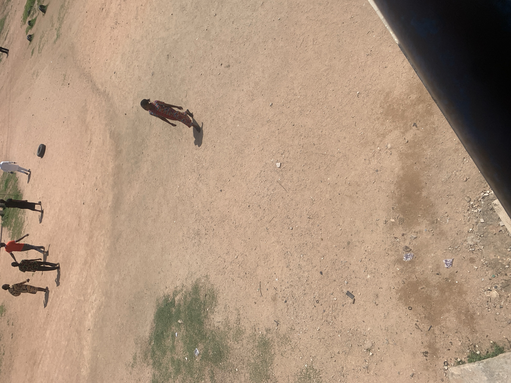 The image shows an aerial view of a dirt ground with several people scattered across it. In the center of the frame, a person in a patterned dress is walking. To the left, four individuals are standing, with one in a red shirt and others in darker clothing. Another person in a white outfit is visible further to the left, near a black tire. The scene appears to be outdoors, possibly a sports field or an open area in a settlement. The lighting suggests it is daytime, with shadows cast by the individuals, indicating the sun is up. The ground is dusty and dry, with patches of sparse green vegetation visible. Some debris, including pieces of plastic and what looks like candy wrappers, are scattered on the ground, particularly in the lower right section. The overall impression is a casual gathering or activity taking place in a somewhat unkempt outdoor environment.