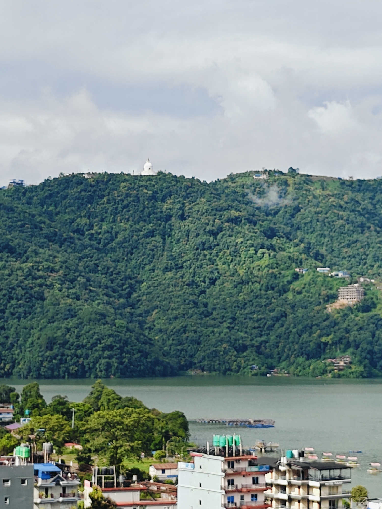This is a wide shot of a lake in Pokhara, Nepal. On the left side of the frame, there is a cluster of buildings, some of which are residential and some appear to be hotels. In the foreground, there are a few more buildings and some vegetation. The lake is in the middle ground, with a few small boats and floating structures. In the background, a large, tree-covered mountain rises, with a white stupa visible at the summit. The sky is overcast.