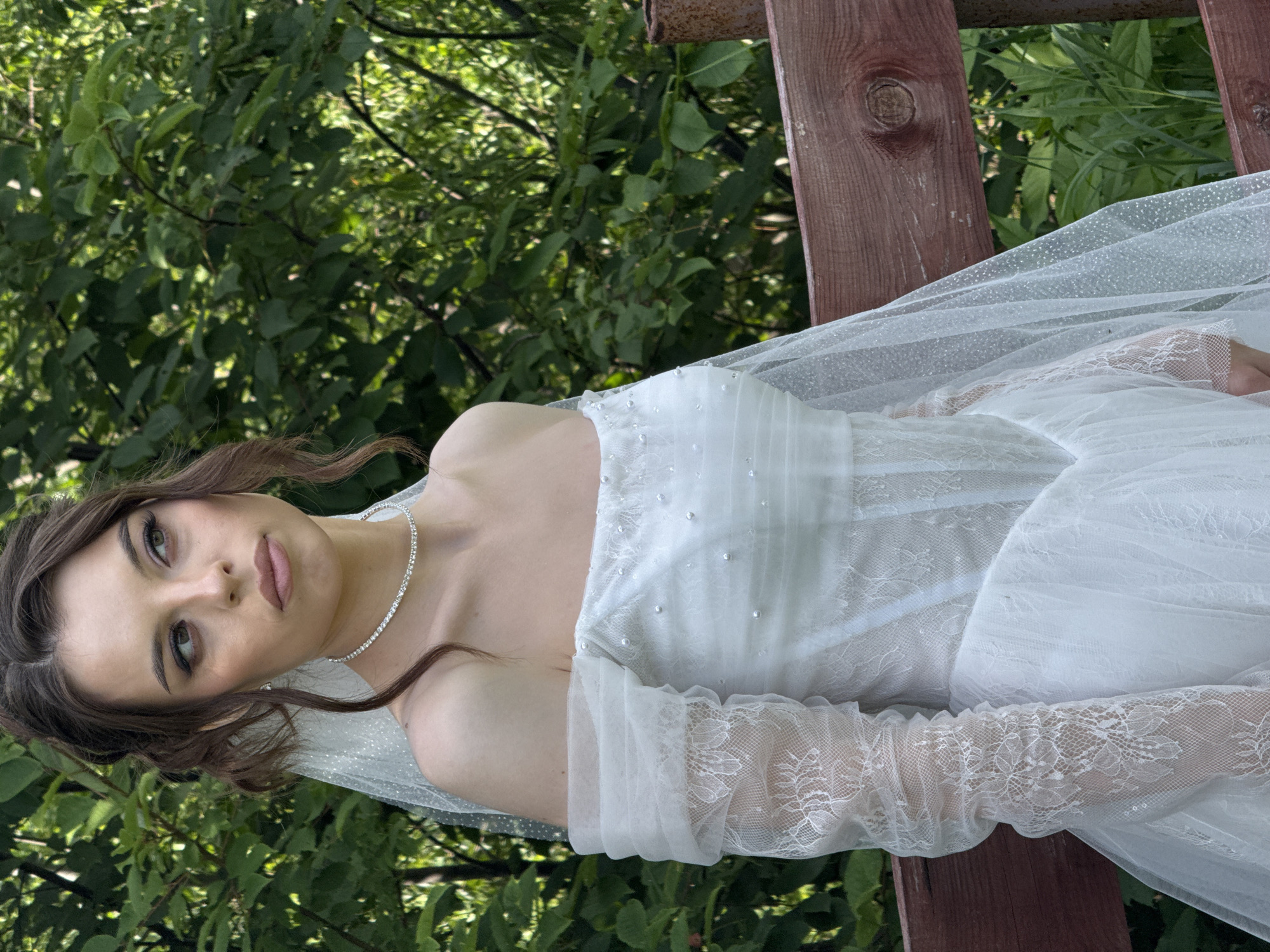 A young woman, dressed in a white wedding gown with off-the-shoulder sleeves and a sheer veil adorned with sparkles, stands outdoors. The wedding dress features intricate lace detailing on the sleeves and is embellished with small pearls across the bodice. She wears a delicate diamond necklace. Her dark hair is styled up with loose tendrils framing her face, and she has on noticeable makeup, including winged eyeliner and lip color.
The woman is positioned in front of a dense backdrop of green foliage, suggesting a natural or garden setting. To her right, a weathered wooden fence runs horizontally across the frame, with a rusty metal post visible at the right edge. The lighting indicates it is daytime, with soft illumination casting gentle shadows.
The location is identified as Votkinsk, Russia. The scene appears to capture a moment of a wedding photoshoot or celebration.
