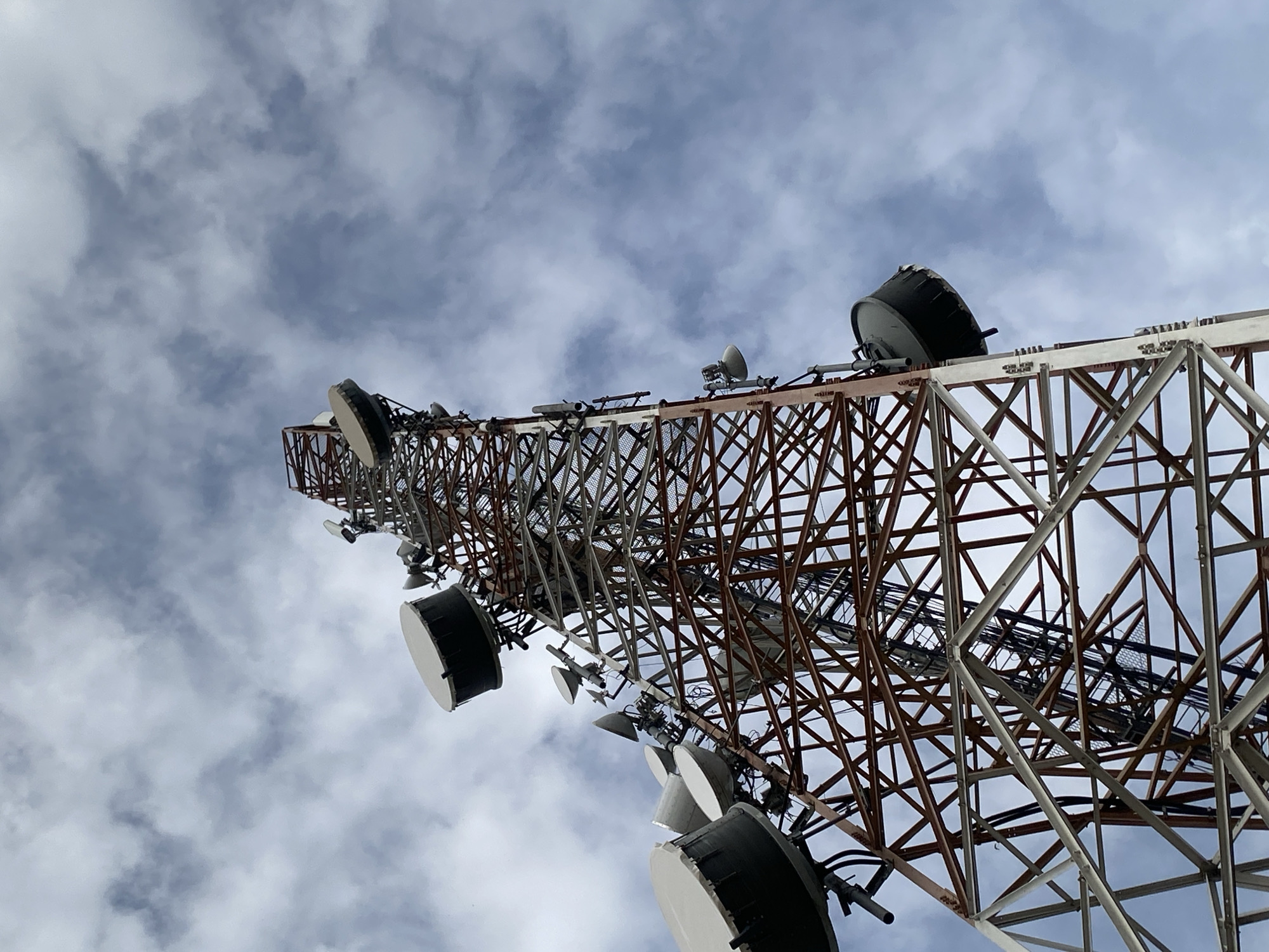 This low-angle, wide shot shows a large telecommunications tower against a cloudy sky. The tower is a lattice structure, with a mix of reddish-brown and white-painted metal beams. Attached to the tower at various levels are numerous satellite dishes, varying in size and shape, with most being round and white or gray. Some dishes are angled upwards, while others are pointed outwards. The sky is filled with a mix of white and gray clouds, suggesting it may be daytime, possibly overcast or with intermittent sun. The perspective looking upwards emphasizes the immense scale of the tower. No people are visible in the frame, nor is any activity actively taking place beyond the static presence of the tower and its equipment. The overall scene is industrial and functional, suggesting a utilitarian purpose for the structure, likely related to communication or broadcasting services in Keffi, Nigeria. No visible text is discernible on the tower or its components.