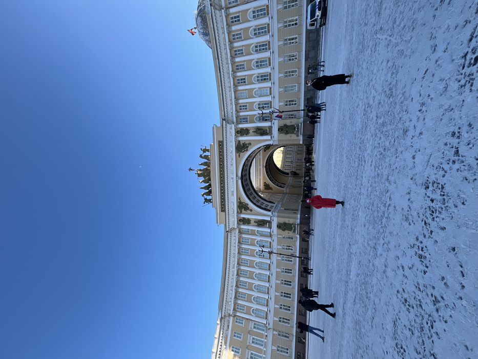 The image presents a wide view of Palace Square in Saint Petersburg, Russia, under a clear, bright blue sky, with a faint crescent moon visible in the upper right quadrant. The ground is covered by a layer of textured white snow, marked with numerous footprints indicating recent pedestrian traffic.
The dominant architectural feature is the General Staff Building, a large, curving, neoclassical structure with a light cream facade, numerous windows, and decorative columns and architectural details. Central to this building is a grand triumphal arch, crowned by a bronze statuary group depicting a chariot drawn by six horses, driven by a figure, flanked by additional figures, commonly known as the Chariot of Glory. Through the arch, further architectural elements of the city are visible, illuminated by natural light. On the far right side of the building, a large dome structure is visible, with a red flag, likely Russian, flying from its apex. Several Russian flags are also affixed to poles on the building's facade near the arch.
Multiple individuals are present in the square. In the mid-foreground, a person wearing a bright red long coat stands centrally, facing generally towards the viewer. To their right and slightly further back, another person in a dark, long coat stands with their left side partially visible, facing right. To the left, an individual in dark clothing is captured mid-stride, walking from left to right across the snow. Numerous other people, dressed in winter attire, are scattered across the square and gathered near the base of the General Staff Building, with many appearing to walk towards or through the triumphal arch. A white service van with dark markings is parked on the far right edge of the frame, adjacent to the building. A lamppost stands in the mid-ground, to the left of the arch.