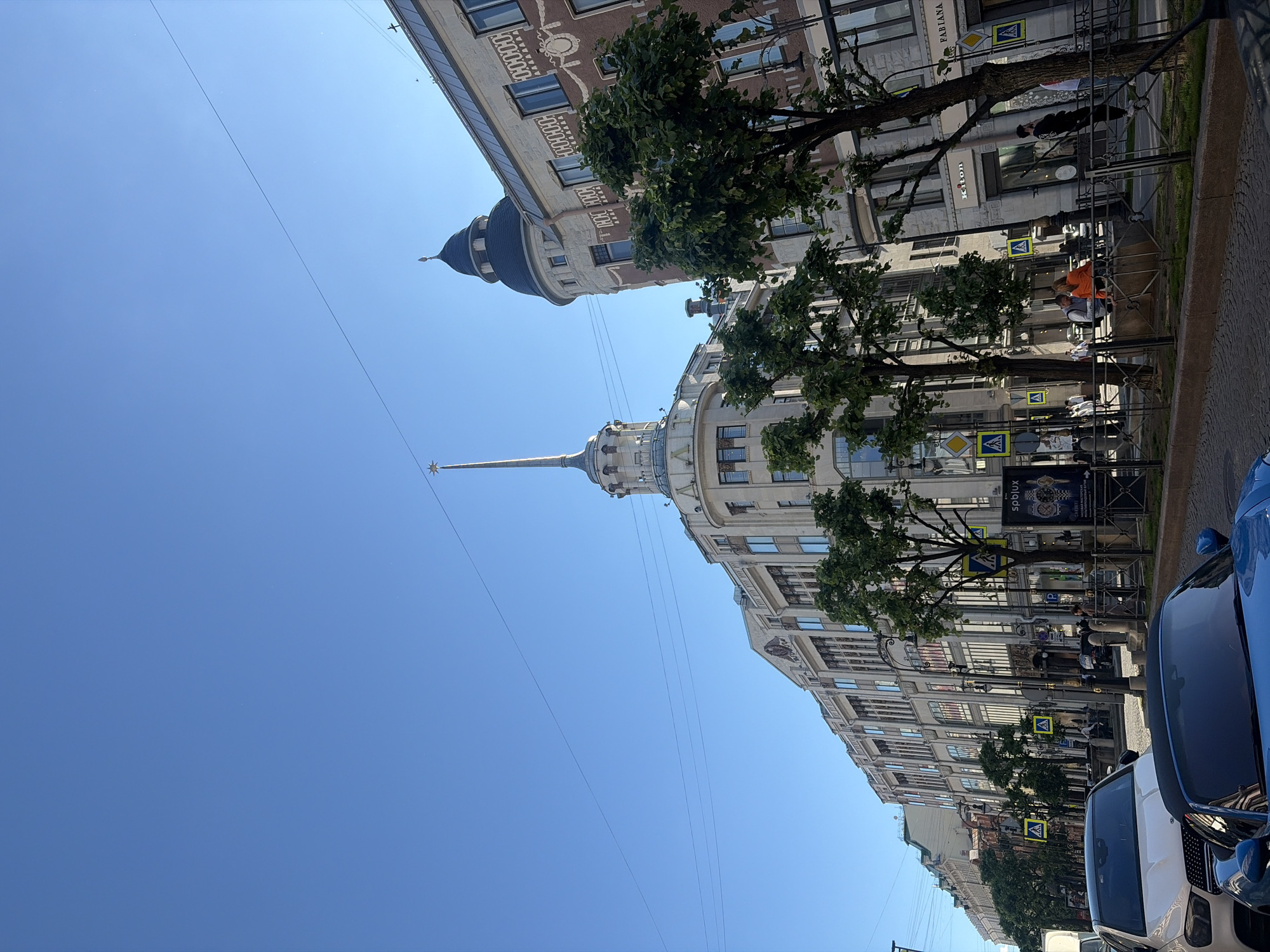 The image captures a street scene in Saint Petersburg, Russia, under a clear blue sky. The perspective is from street level, looking up at the facades of several ornate buildings. On the left, a white SUV is parked next to a blue convertible. The street is paved with cobblestones and lined with trees.
Dominating the scene are the architectural features of the buildings. The building in the center features a distinctive spire topped with a star, a common architectural element in Saint Petersburg. Adjacent to it, to the right, is a building with a dark, rounded tower. The facades of the buildings are richly decorated with classical architectural details, including numerous windows, columns, and patterned stonework.
Several yellow pedestrian crossing signs are visible along the sidewalk. In the middle ground, people are seated on a low stone wall, some of them interacting. Other pedestrians are walking along the sidewalk. A poster advertising watches is displayed on a stand near the sidewalk. The overall atmosphere is that of a sunny day in a historic European city.