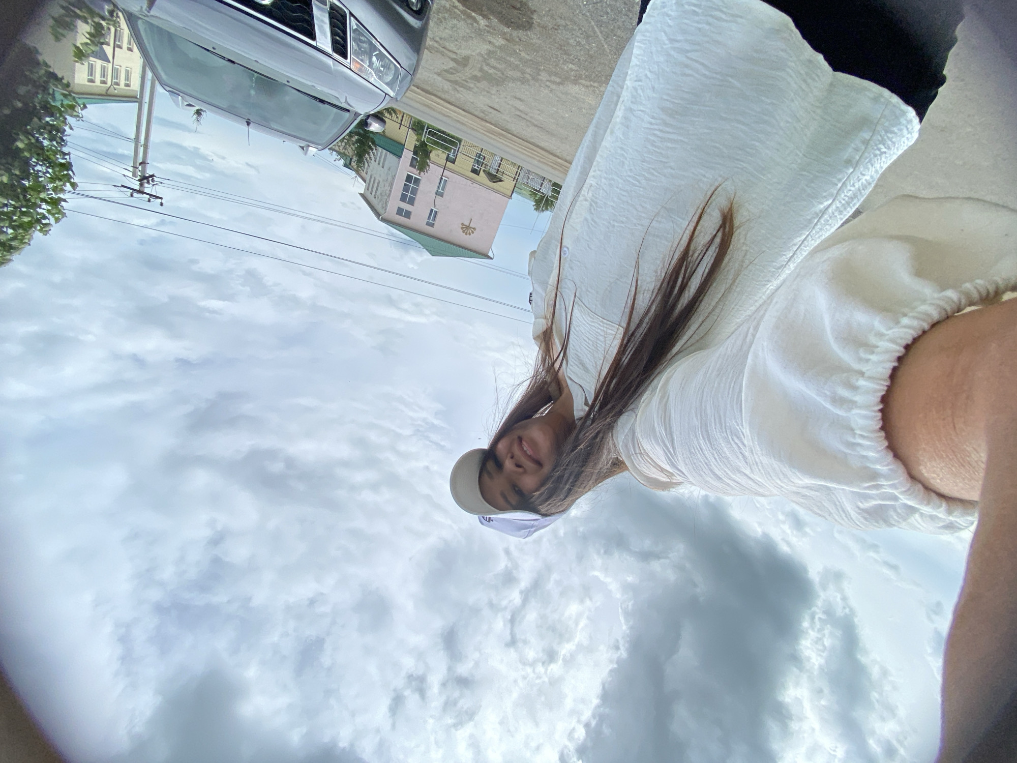 A low-angle, wide-angle selfie taken outdoors on an overcast day shows a young woman from the chest up, facing the camera with a slight smile. She is wearing a white baseball cap and a crinkled white shirt with elastic at the cuffs. Her long, dark hair is visible. In the background, a gray SUV is parked. Beyond the vehicle, a pink and green building with several windows is visible, along with utility poles and wires. The ground appears to be a mixture of asphalt and gravel. The scene is consistent with an outdoor urban environment in Akyar, Russia.