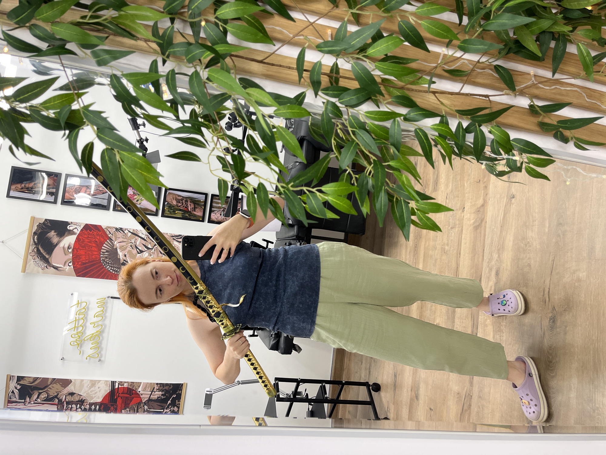 A woman with auburn hair is standing in what appears to be a tattoo parlor, judging by the artwork on the walls and the tattoo chair visible in the background. She is holding a decorative sword with a golden hilt and black sheath. She is wearing a dark blue sleeveless top and loose, light green pants, along with purple clogs. She is taking a selfie in a large mirror. The environment includes a wall with framed artwork and a neon sign, a trolley with tattoo equipment, and a large artificial plant with green leaves. The floor is wooden. The overall lighting suggests an indoor setting.
