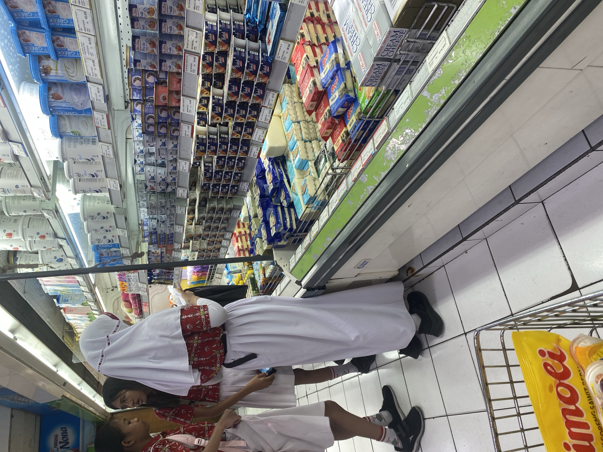 The image depicts three young females in a grocery store, likely a supermarket, in Manado, Indonesia. Two of the individuals are wearing school uniforms consisting of red tops with white collars and white skirts. The third individual, standing slightly behind them, is wearing a white headscarf and a long white skirt, also with a red-patterned top. They appear to be looking at products displayed on refrigerated shelves filled with dairy items, including yogurt and cheese. The shelves are stocked with various brands and flavors of yogurt in tubs and cheese in blocks and packages. In the foreground, a shopping cart is partially visible, containing a yellow package with Indonesian text, possibly snacks or food items. The store environment features tiled flooring and overhead fluorescent lighting, consistent with a retail setting. The overall scene suggests a casual shopping trip or an outing after school.