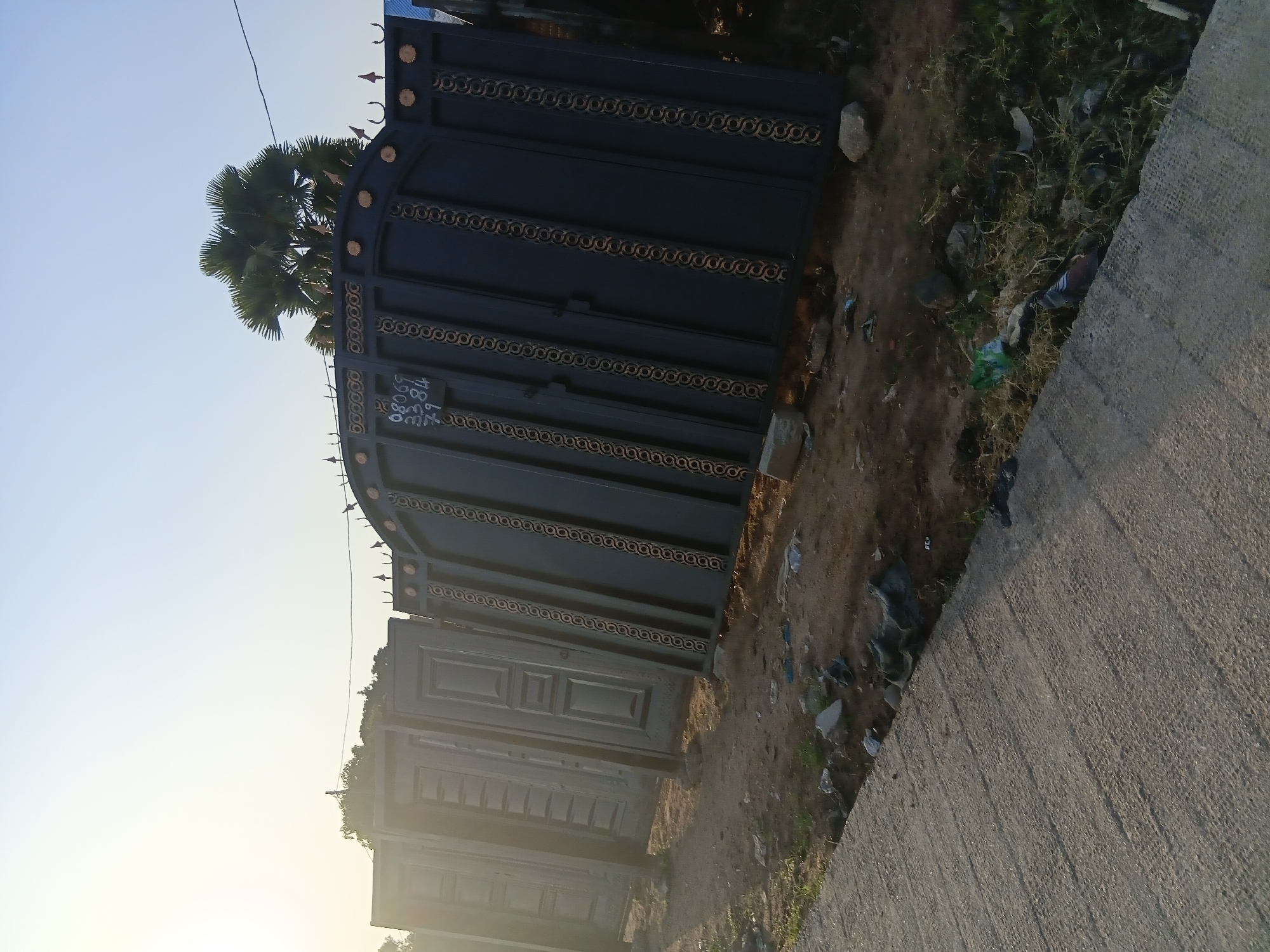 The image shows a street scene in Jalingo, Nigeria, with several large, dark metal gates leaning against each other in the foreground. The gates are ornately decorated with gold trim and have a spiked design at the top. To the left of the gates, what appear to be multiple doors are also leaning, displaying panel designs. The ground is dirt and debris, with some grass and scattered trash. A paved walkway or road is visible in the lower left corner of the image. In the background, a palm tree stands against a bright, hazy sky, with electrical wires strung overhead. The scene appears to be an outdoor display or storage area for the gates and doors.
