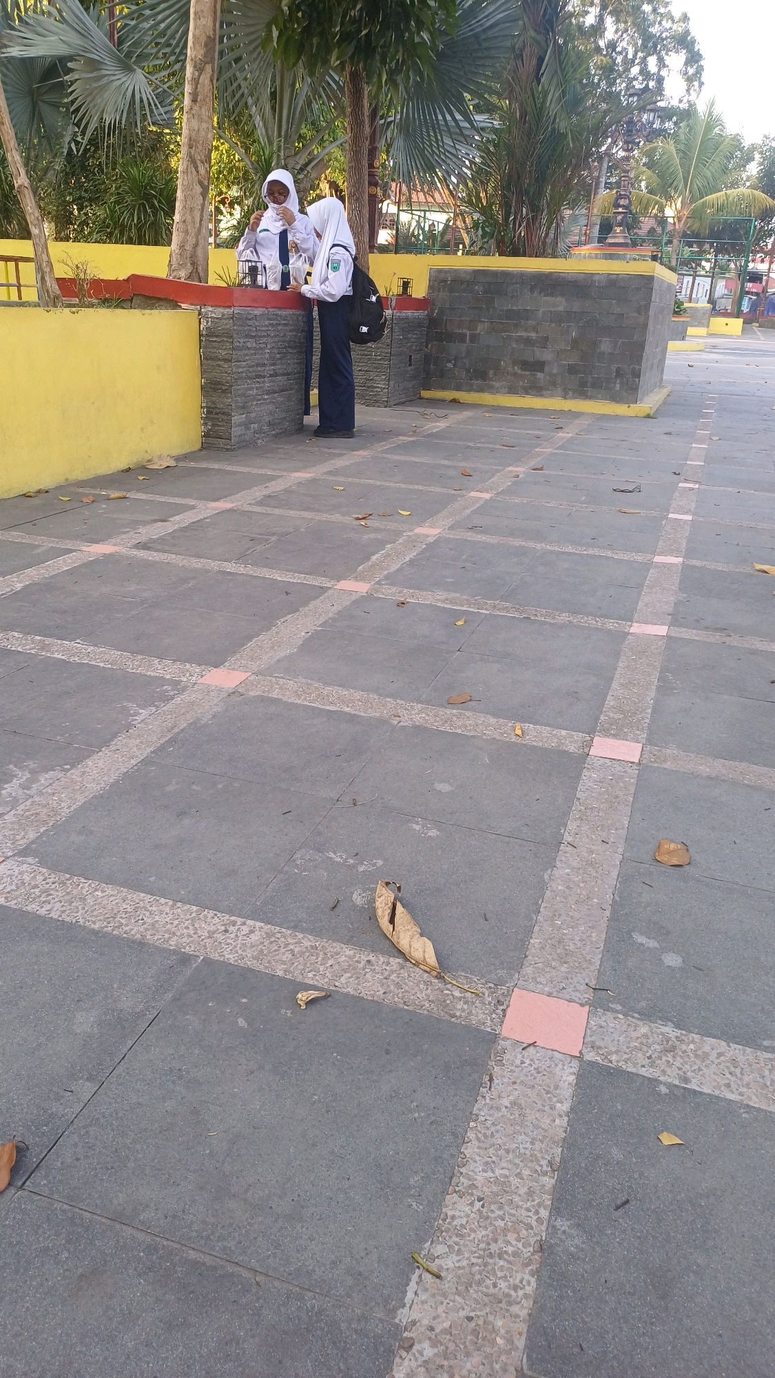 The image shows two young females in school uniforms, one in a white hijab and the other with a white hijab and uniform. They are standing by a yellow wall and a grey stone structure. One of the students appears to be holding something white and translucent, possibly a drink. The ground is paved with large grey tiles, interspersed with pink squares and a textured border. Scattered leaves are visible on the ground, suggesting it might be autumn. In the background, there are trees with large fronds and a glimpse of buildings. The lighting indicates it is daytime, with sunlight casting shadows. The setting appears to be an outdoor public space, possibly a schoolyard or park area within Klaten, Indonesia.