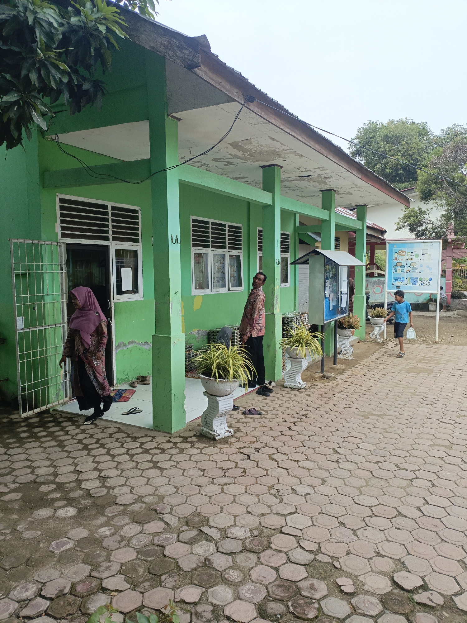 The image depicts the exterior of a one-story building with a green facade and a weathered, off-white overhang. The building appears to be a public facility, possibly a school, given the presence of windows with metal bars and a bulletin board displayed outside. The ground is paved with hexagonal interlocking bricks.
In the foreground, a woman wearing a pink hijab and a patterned dress is partially visible through a metal gate, stepping out of a doorway. To the right, a man in a patterned shirt and dark trousers stands by the building. Further to the right, a young boy in a blue t-shirt and dark shorts is walking away from the viewer, carrying a white plastic bag. Two potted plants in ornate white planters are positioned along the building's facade, one near the doorway where the woman is, and another further down. A metal information stand is also present next to the building.
In the background, trees and other buildings can be seen, suggesting an urban or semi-urban environment. The sky is overcast. The overall condition of the building and its surroundings suggests a functional but somewhat aged facility. Based on the context provided, this scene is located in Lhokseumawe, Indonesia.