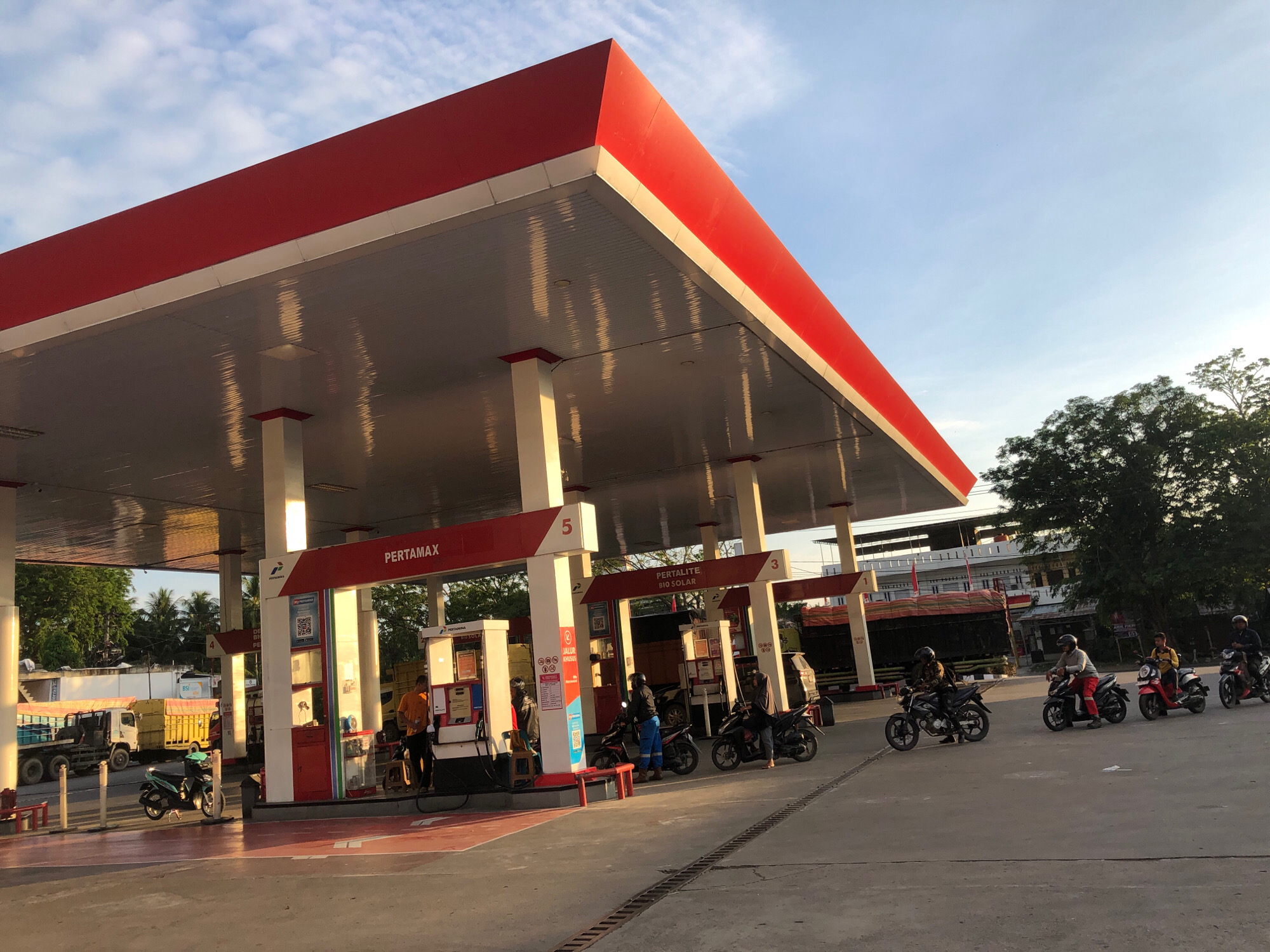 A Pertamina gas station in Lhokseumawe, Indonesia, is depicted under a bright, partly cloudy sky. The main structure features a prominent red roof with white trim, supported by white pillars. Several gas pumps are visible, with one displaying the "PERTAMAX" logo.
A man in an orange shirt stands at a gas pump, attending to a motorcycle. Another man in blue pants and a helmet is seated on a motorcycle near another pump. To the right, several people on motorcycles are gathered, with one person appearing to refuel. Further down the line of pumps, two more individuals on motorcycles are present.
In the background, several trucks are parked, including a yellow truck with a blue bed and a dark-colored truck. The environment includes trees and buildings in the distance, suggesting an urban or semi-urban setting. The pavement is a light gray concrete, and there are some visible drain grates. The overall scene appears to be during daylight, with sunlight casting shadows.