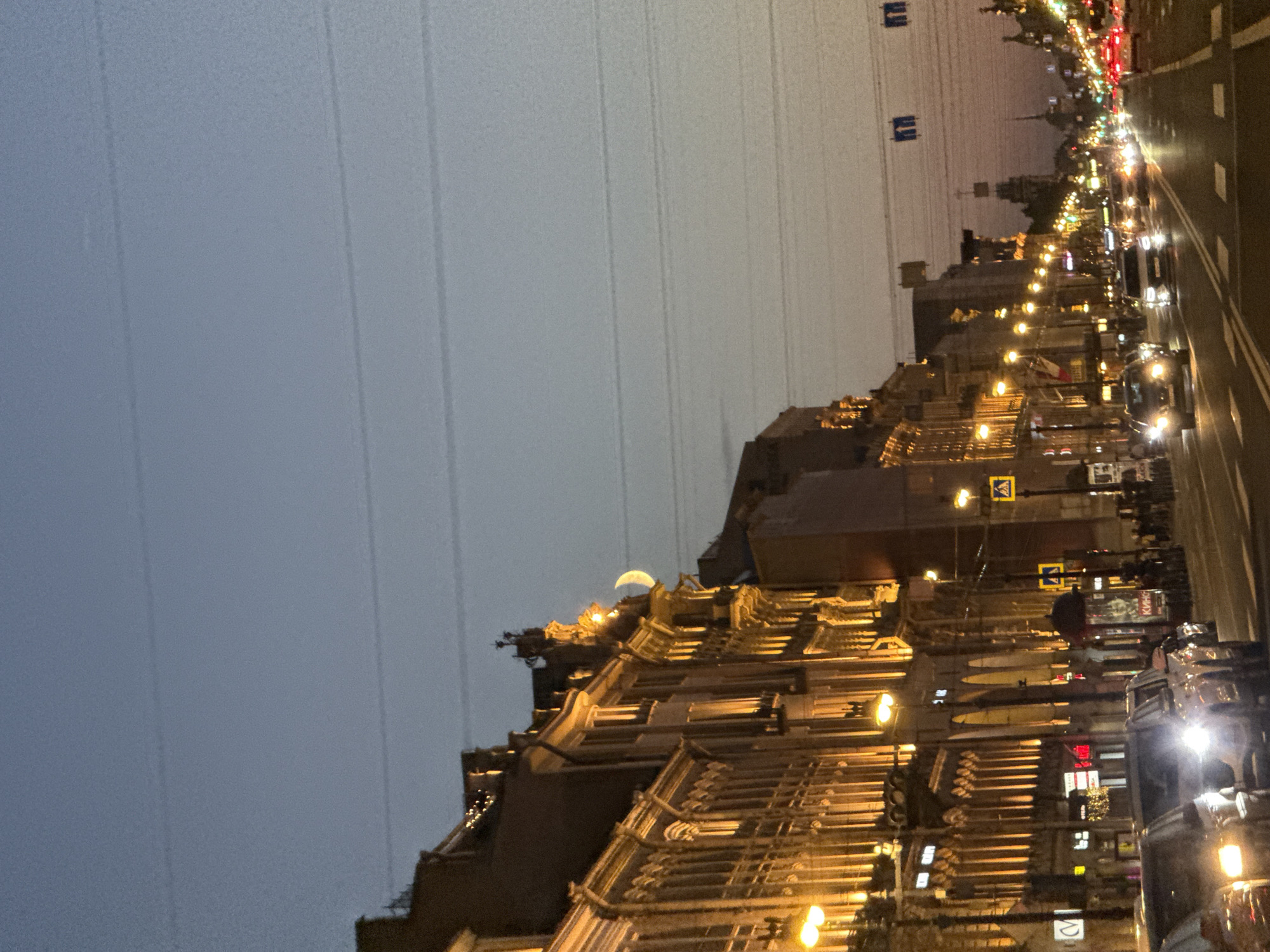 The image depicts a street scene in Saint Petersburg, Russia, at dusk. Ornate, historic buildings line the left side of the street, illuminated by warm, artificial lights, highlighting their detailed architecture. Multiple vehicles, including cars and possibly a bus, are visible on the road, with headlights on. The sky above is a gradient of blues and grays, with faint horizontal lines that could be power lines or contrails. A sliver of the moon is visible against the sky, positioned above the buildings. Traffic lights and street signs are present, indicating an urban environment. Some pedestrians can be seen on the sidewalks. The overall impression is that of a bustling city street transitioning from day to night.