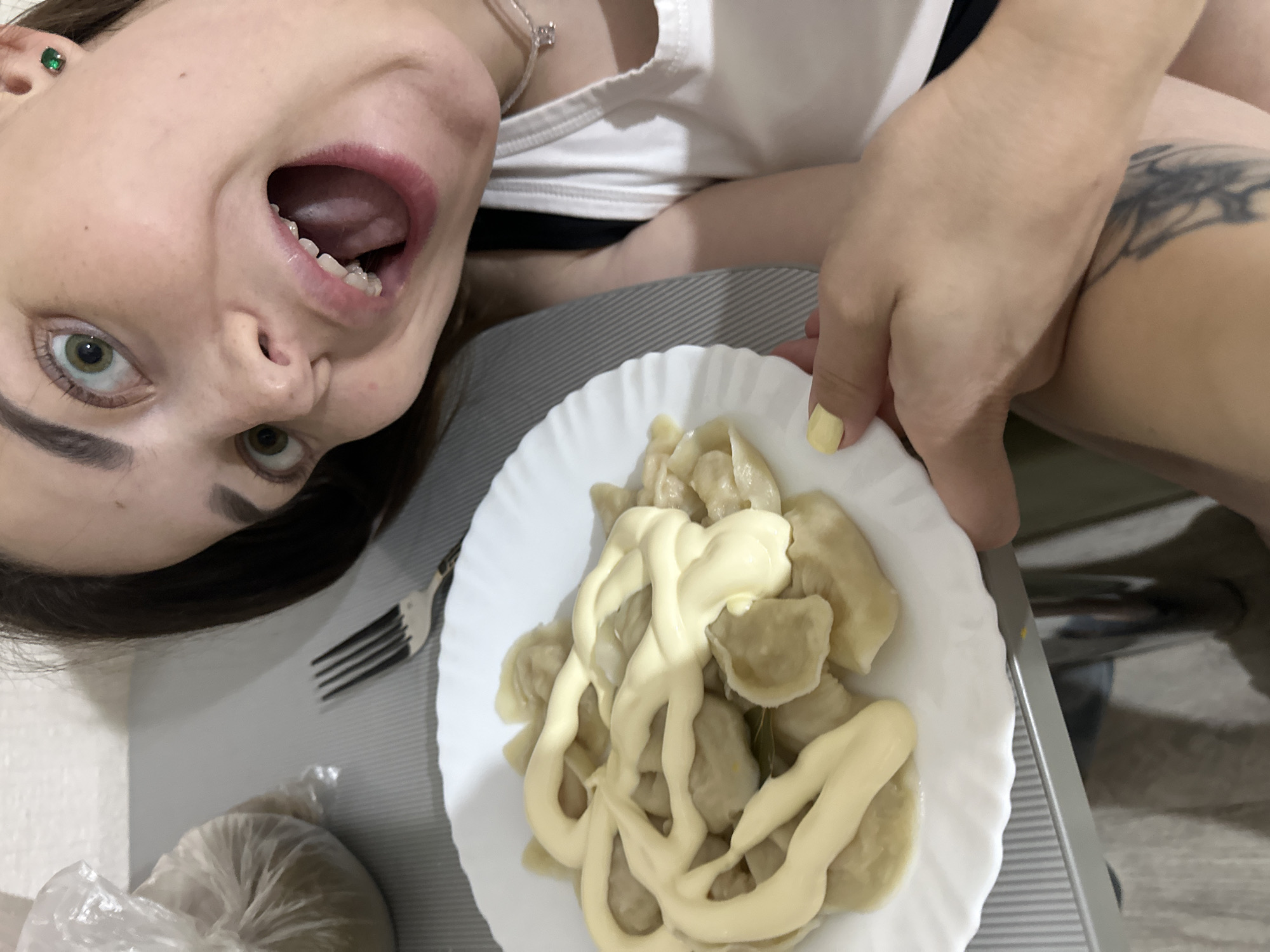 A close-up, waist-up shot shows a young woman with wide eyes and an open mouth, her face angled towards the camera. She has dark hair and is wearing a white tank top and a necklace with a small charm. Her left hand, with a yellow nail, is holding a white plate filled with pierogi topped with a creamy sauce. In the background, to the left, is a plastic bag of what appears to be dough. A fork rests on the table next to the plate. The scene is set indoors, on a table with a grey, striped surface. There is no visible indication of the location being in Perm, Russia.