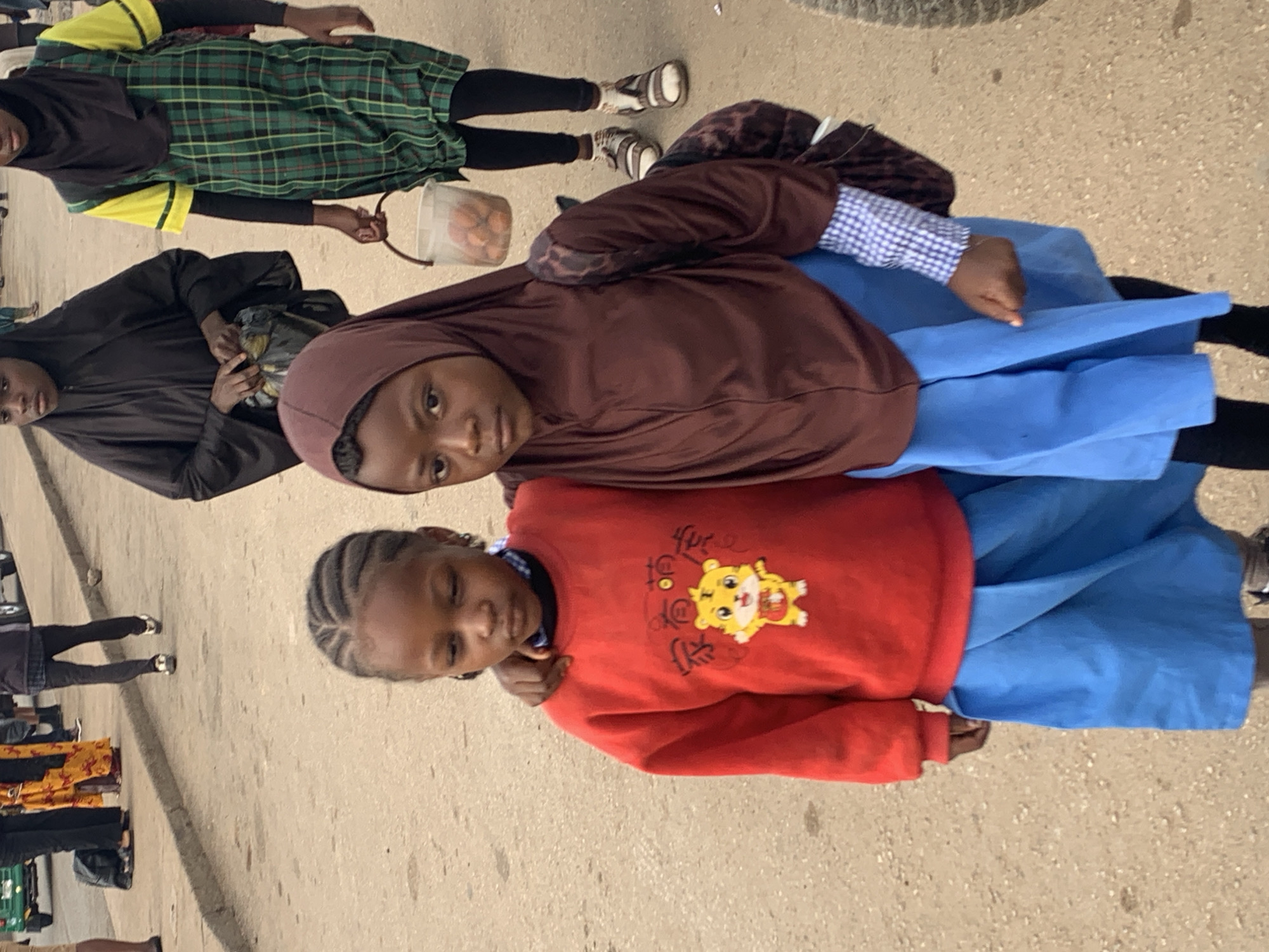Two young girls, likely school-aged, are walking side-by-side on a dusty, paved street. The girl on the left wears a red sweatshirt with a cartoon tiger graphic and a blue skirt. Her hair is braided. The girl on the right wears a brown hijab and a blue skirt, with a checkered shirt visible under her hijab. The girl on the right has a backpack slung over her shoulder. In the background, other individuals are visible walking along the street. One person on the right is wearing a green and yellow plaid dress over black leggings and sandals, carrying a white bucket containing small, round objects. The environment appears to be a typical street scene in a Nigerian city, possibly Gura Top, characterized by the paved road, dusty ground, and casually dressed pedestrians.