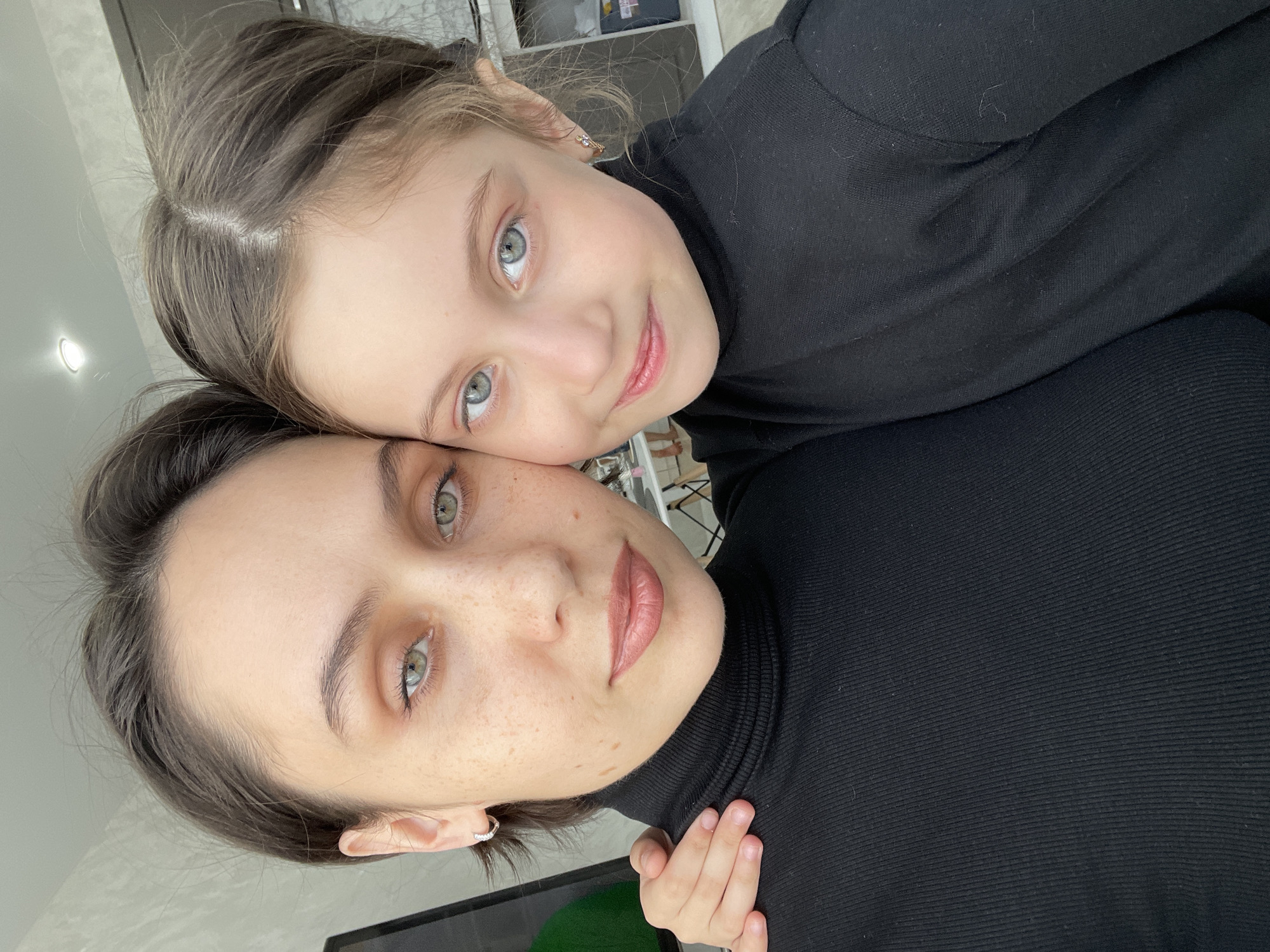A selfie shows a woman and a child, likely a daughter, posing closely together. Both are wearing black turtleneck shirts. The woman is in the foreground with the child nestled behind her, resting her head against the woman's. Both are looking at the camera with soft smiles. The background is blurred, showing a room with neutral tones. No evidence suggests this image was taken in Izhevsk, Russia.