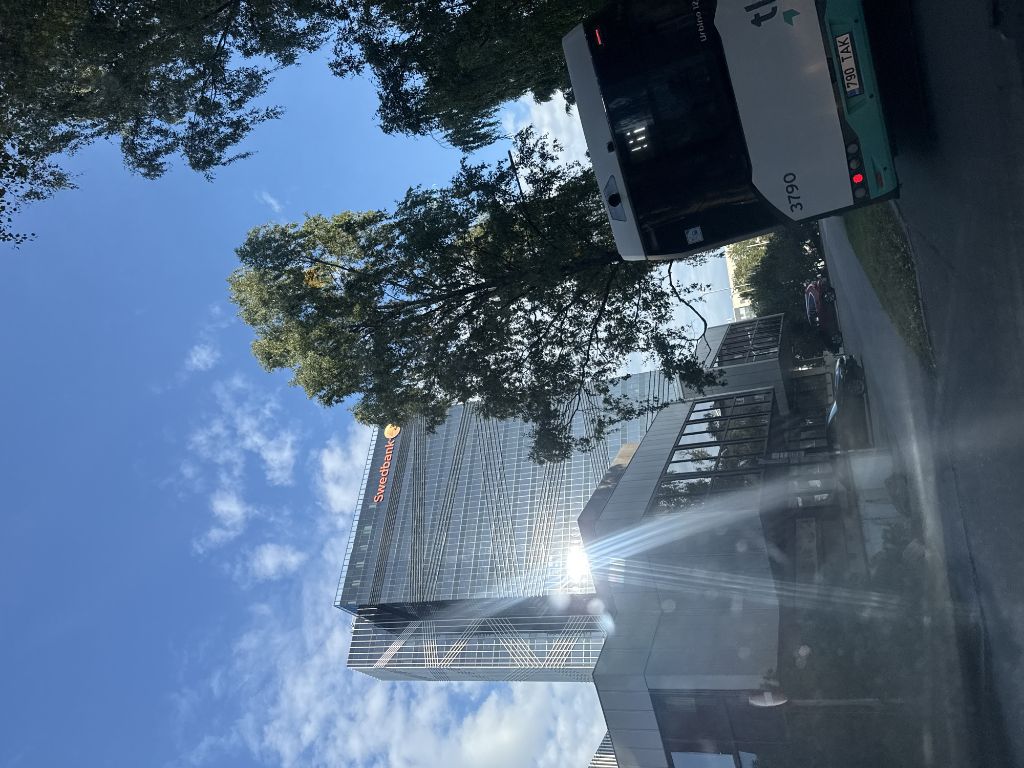 The image captures a view of the Swedbank building in Tallinn, Estonia, on a clear day with scattered clouds. The prominent modern glass skyscraper, with its distinctive reflective facade and "Swedbank" signage in orange, stands tall against the blue sky. In the foreground, a bus is visible from the rear, displaying the number "3790" and license plate "790 TAK". Trees with green foliage frame the scene on the right and partially obscure the sky. The sun is bright, casting strong lens flares and reflections on the Swedbank building and a lower adjacent structure with glass windows. A road is visible in the lower part of the image, with parked cars on the left side. The overall impression is an urban streetscape featuring a notable corporate landmark.