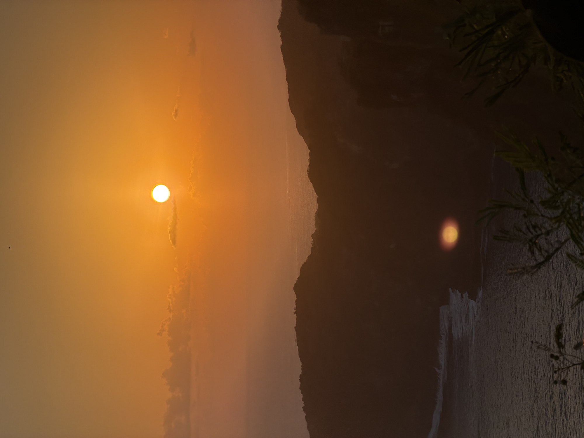 The image depicts a sunset over the ocean in Nusa Penida, Indonesia. The sun, a bright, orb, is positioned in the upper center of the frame, casting a warm orange glow across the sky and a hazy, soft light on the landscape below. Scattered, wispy clouds are visible in the sky, some catching the sunlight. In the mid-ground, dark, silhouetted hills or cliffs dominate the scene, their contours softened by the low light. The ocean stretches out before these landforms, appearing as a dark expanse with subtle white lines indicating waves breaking near the shore. In the foreground, the dark shapes of vegetation, likely trees or bushes, are visible, adding depth to the composition. A faint reflection of the sun can be seen on the water's surface. There are no people or discernible human activity in the image.