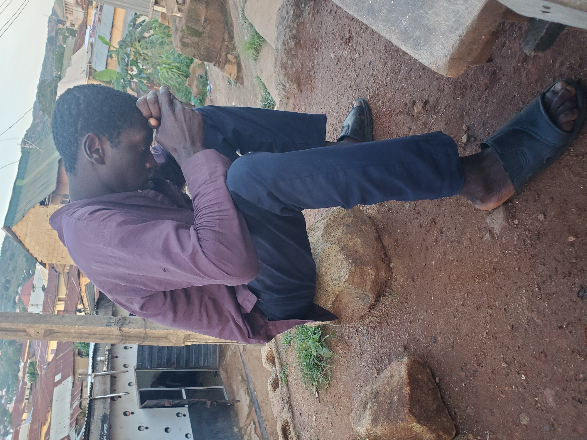 A young Black man is sitting on a rock outdoors, with his head bowed and hands clasped in front of his forehead, suggesting contemplation or distress. He is wearing a dark purple collared shirt and dark blue pants, with black sandals on his feet. The ground is a mix of red dirt and rocks. In the background, there are buildings with corrugated metal roofs, characteristic of a rural or developing area. Some greenery is visible, including bushes and trees. The overall scene appears to be in a rural setting in Gwong, Nigeria. The lighting suggests it is daytime, possibly late afternoon given the angle of the light. No text is visible in the image.