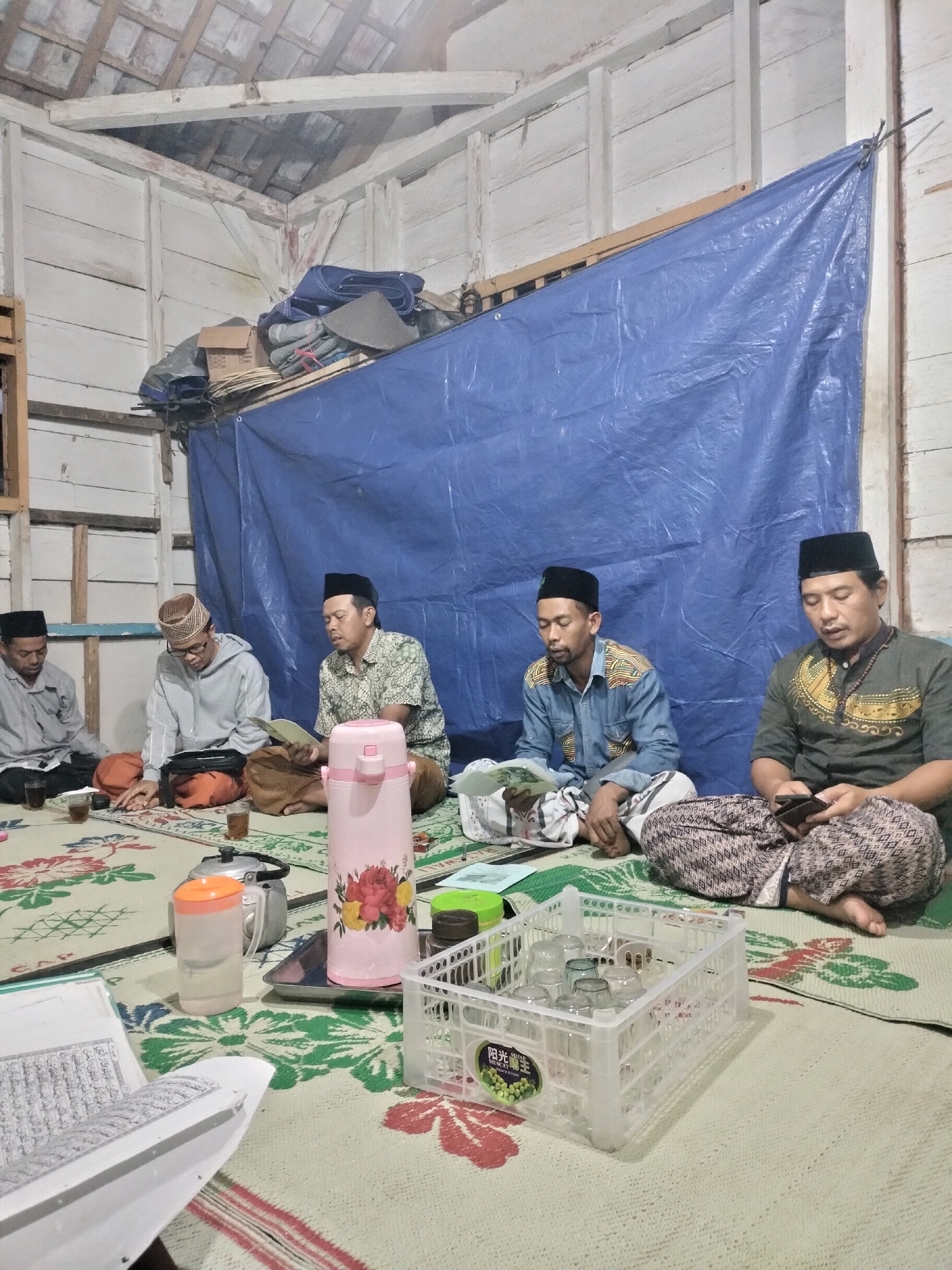 The image depicts a gathering of five adult males seated on mats on the floor in what appears to be an indoor space. The room has white wooden walls and a visible wooden beam ceiling. A large blue tarp is suspended behind the individuals, obscuring the upper portion of the back wall.
The individuals are dressed in traditional Muslim attire, including peci (caps) and sarongs. From left to right, the first man is wearing a grey long-sleeved shirt and dark peci. The second man is wearing a light grey hooded sweatshirt and glasses, with a patterned cap. The third man is in a patterned light-colored shirt and a dark peci. The fourth man is wearing a blue denim-like shirt with patterned shoulders and a dark peci. The fifth man, on the far right, is wearing a dark green shirt with gold patterned accents and a dark peci. He is holding a mobile phone.
Several objects are present on the mats, including tea glasses, a teapot, a pink thermal carafe with a floral design, a pitcher of water, a book open on the floor with Arabic script, and a white plastic basket filled with glasses. The overall atmosphere suggests a religious or communal gathering. The lighting is dim, indicating it is likely evening or indoors without strong natural light. The location is identified as Pujut, Indonesia.