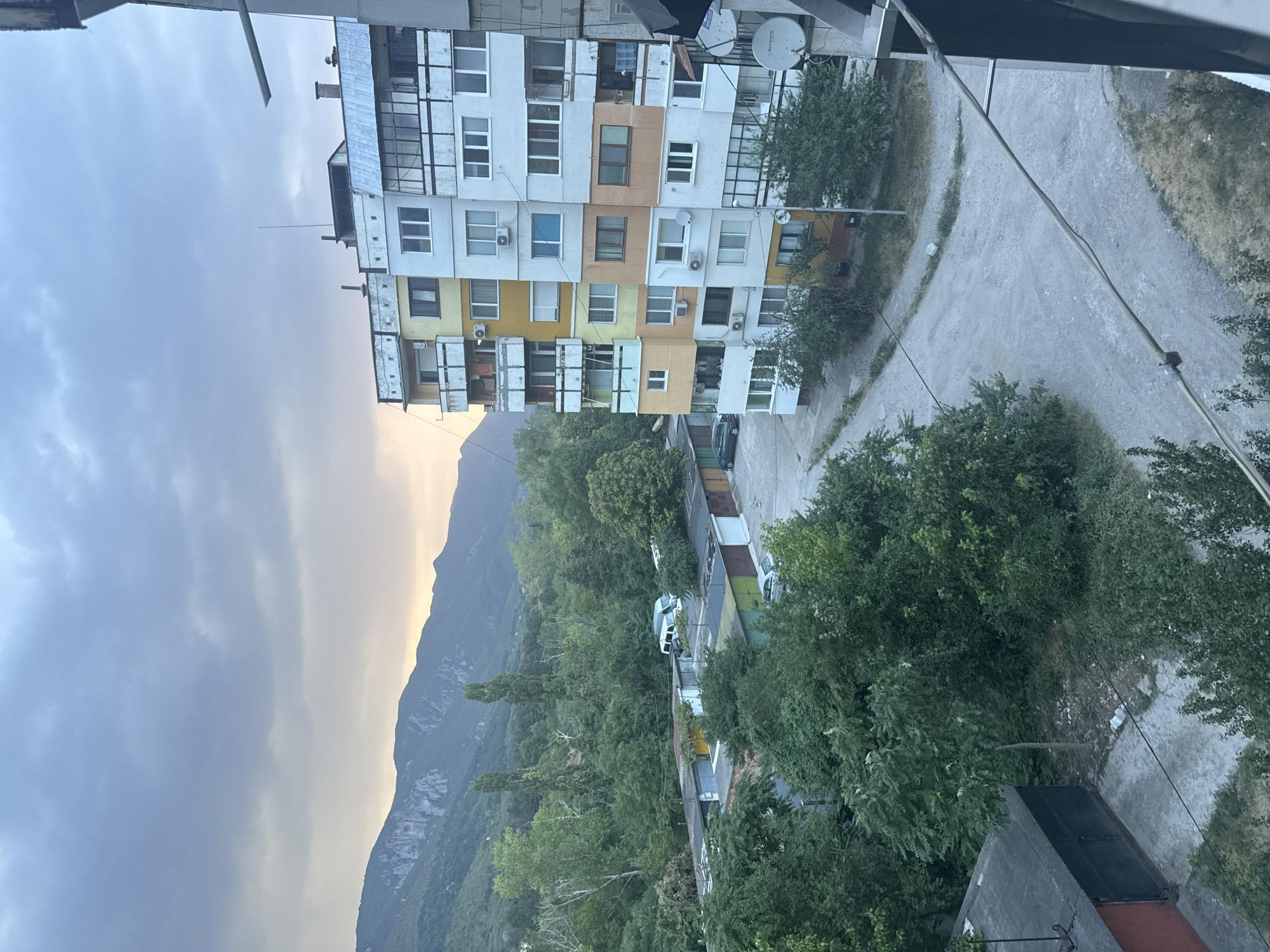 The image is a high-angle, medium shot of a residential area at dusk. The sky is overcast with a gradient of grey and a hint of orange near the horizon, suggesting the sun has recently set. In the foreground, on the right, part of a building's balcony railing is visible, with satellite dishes attached to the exterior wall. To the left, a multi-story apartment building with a colorful facade of yellow, white, and orange blocks dominates the mid-ground. It features numerous windows and balconies, some with laundry hanging out. Below the apartment building is a street or paved area. Further back, a row of garages with different colored doors is visible, interspersed with dense green trees. A white van and a silver car are parked near the garages. In the background, a range of mountains covered in green foliage stretches across the horizon, with rocky outcrops visible on the peaks. The overall scene depicts a somewhat weathered urban environment with a natural backdrop.